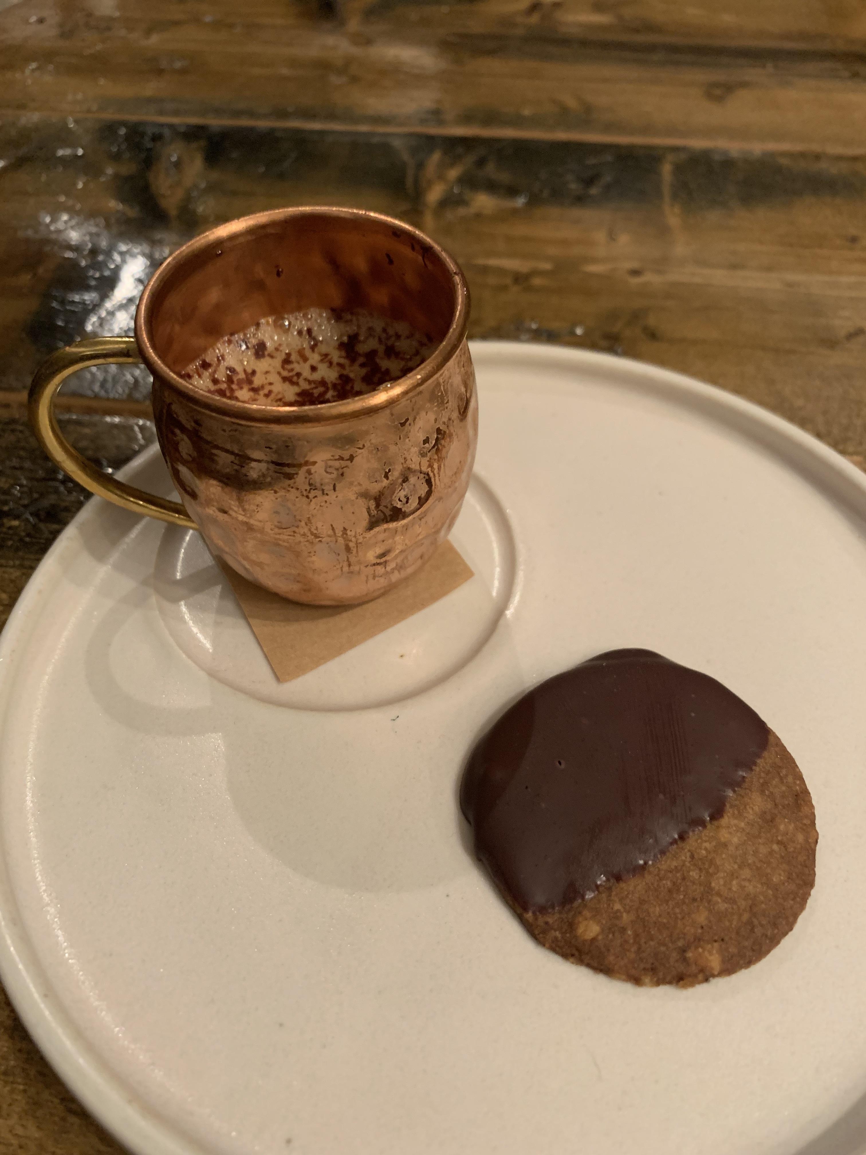 A small cup of milk topped with spices next to a small cookie that has been dipped in chocolate
