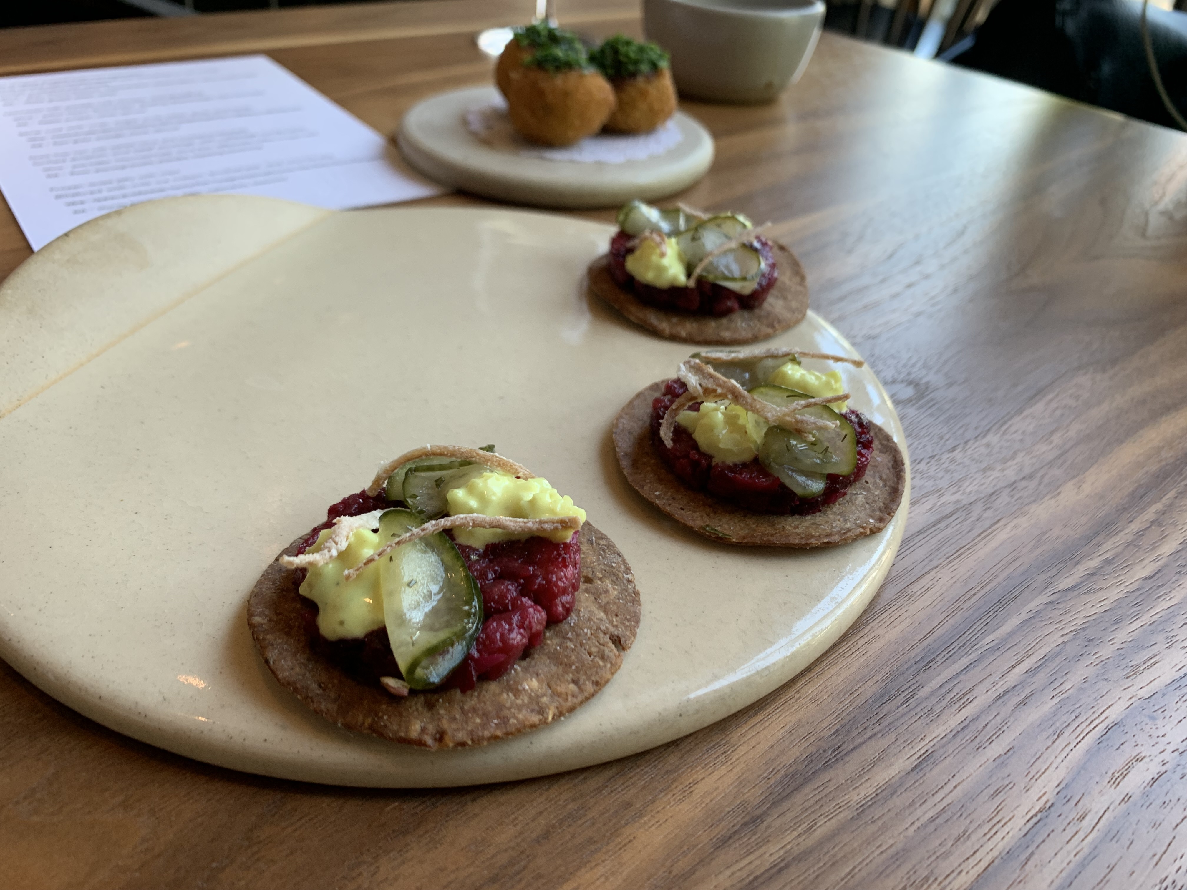 Thin pickles folded over atop a bed of red, raw meat, served on a dark brown disc of cracker.