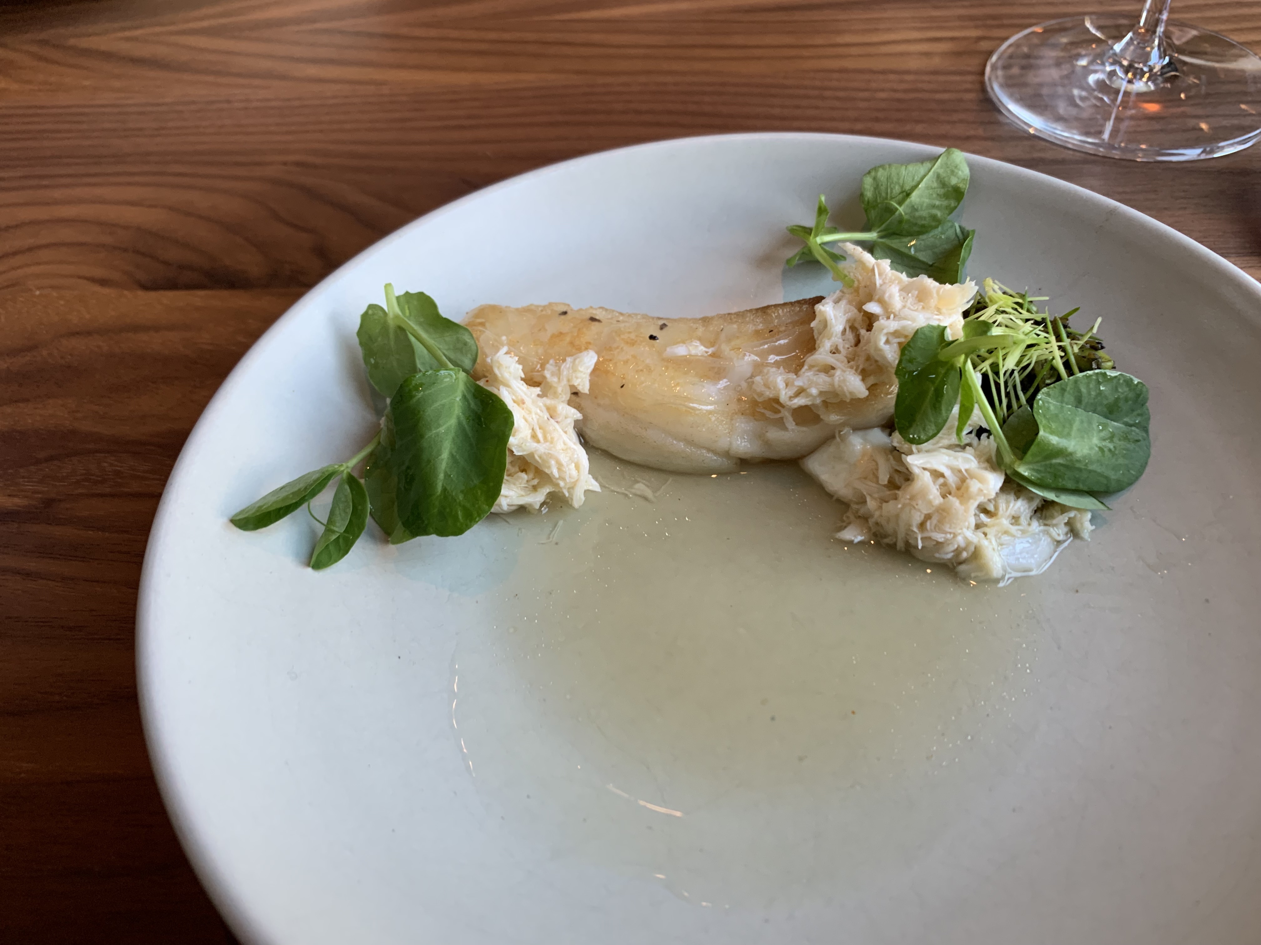 Small cod fillet at the top of a plate, with mounds of crab meat around it, and a few greens accenting it. Directly beneath the filet is a small pool of sauce. The plate is reminiscent of a log next to a pond.