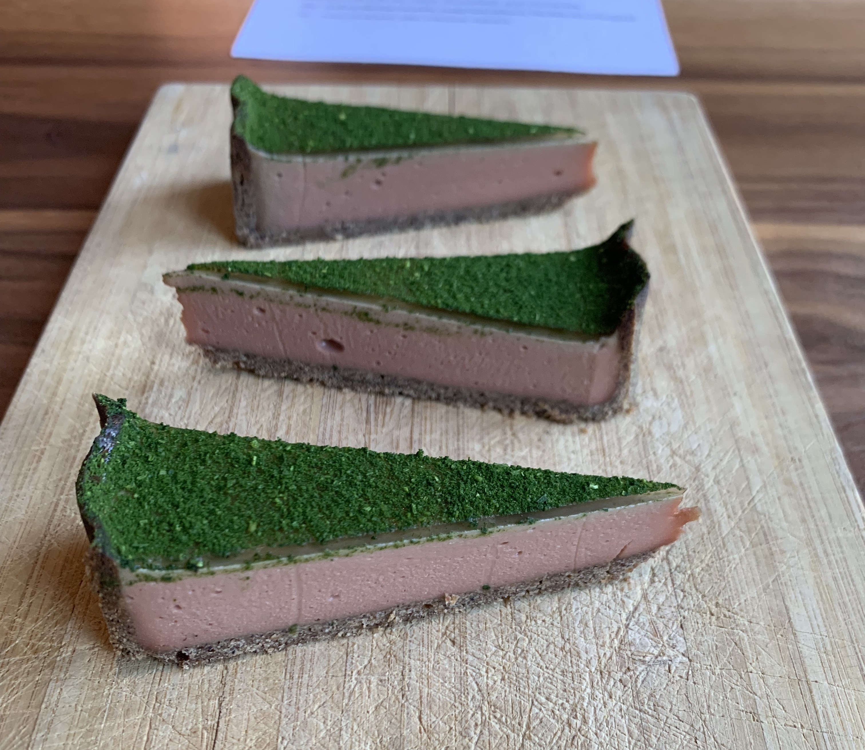 Three slices of tart served on a board, arranged in a tessellated manner. The tart has a topping of fine green powder, and the filling is a fleshy pink colour.