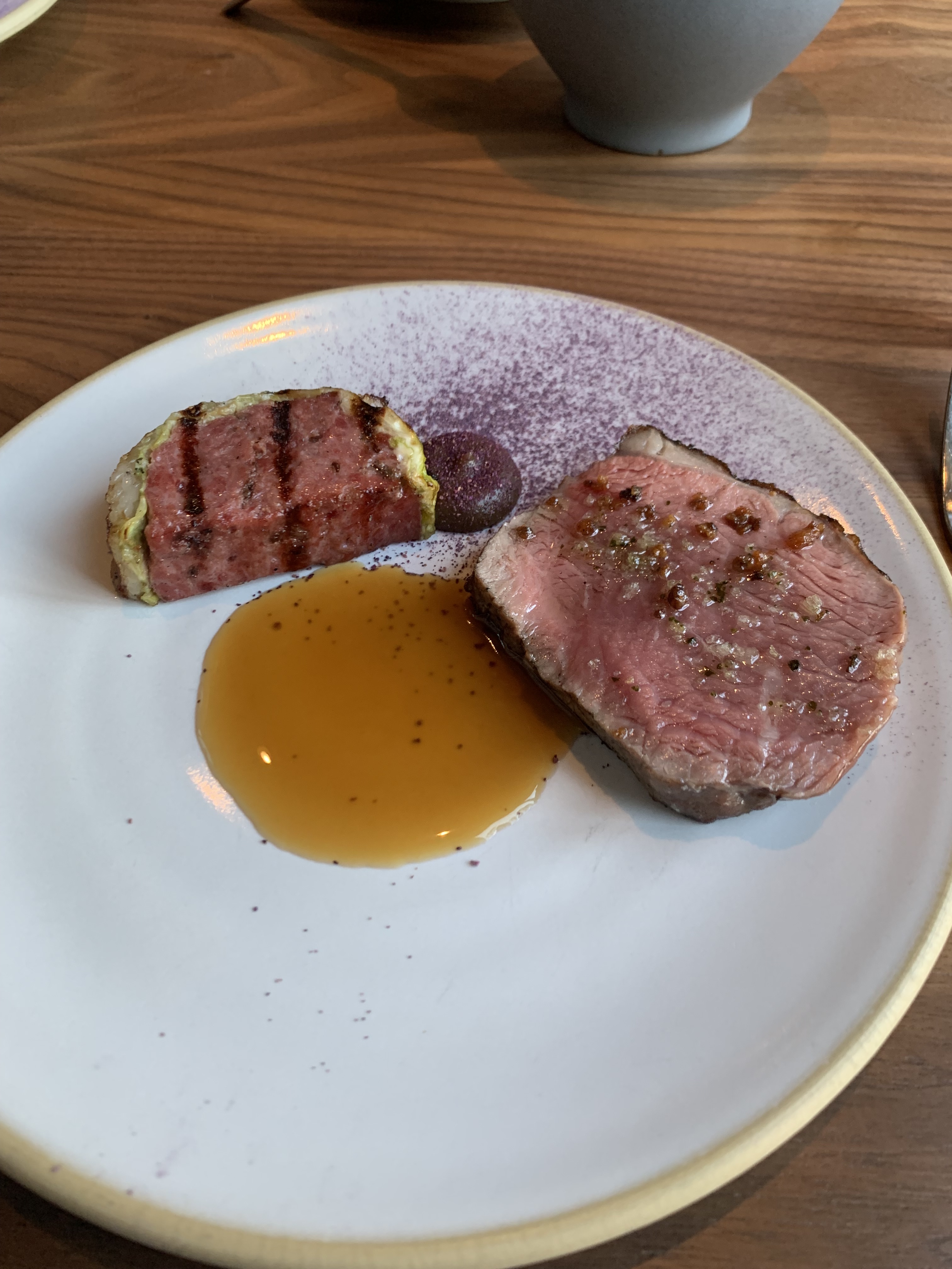 Half-moon slice of sausage wrapped in cabbage, with nice grill marks over it. Served next to a round of medium beef, with a dollop of ketchup between them, and a pool of golden-hued sauce beneath. There is a scattering of purple cabbage dust on the plate for aesthetics. 