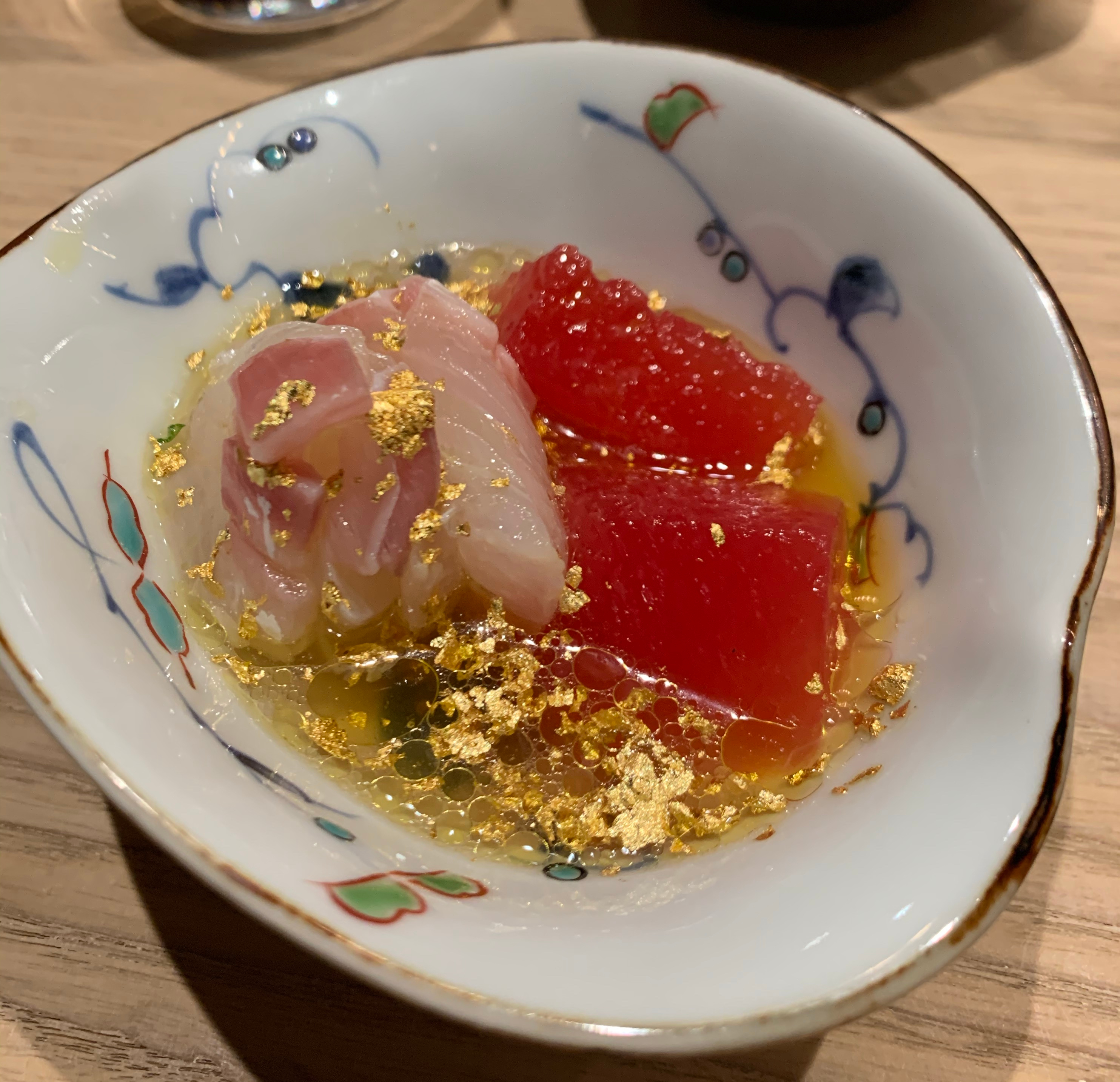 A plate with raw snapper and dark-red watermelon chunks poking out of a light-yellow broth. The whole thing has been covered in gold flakes.
