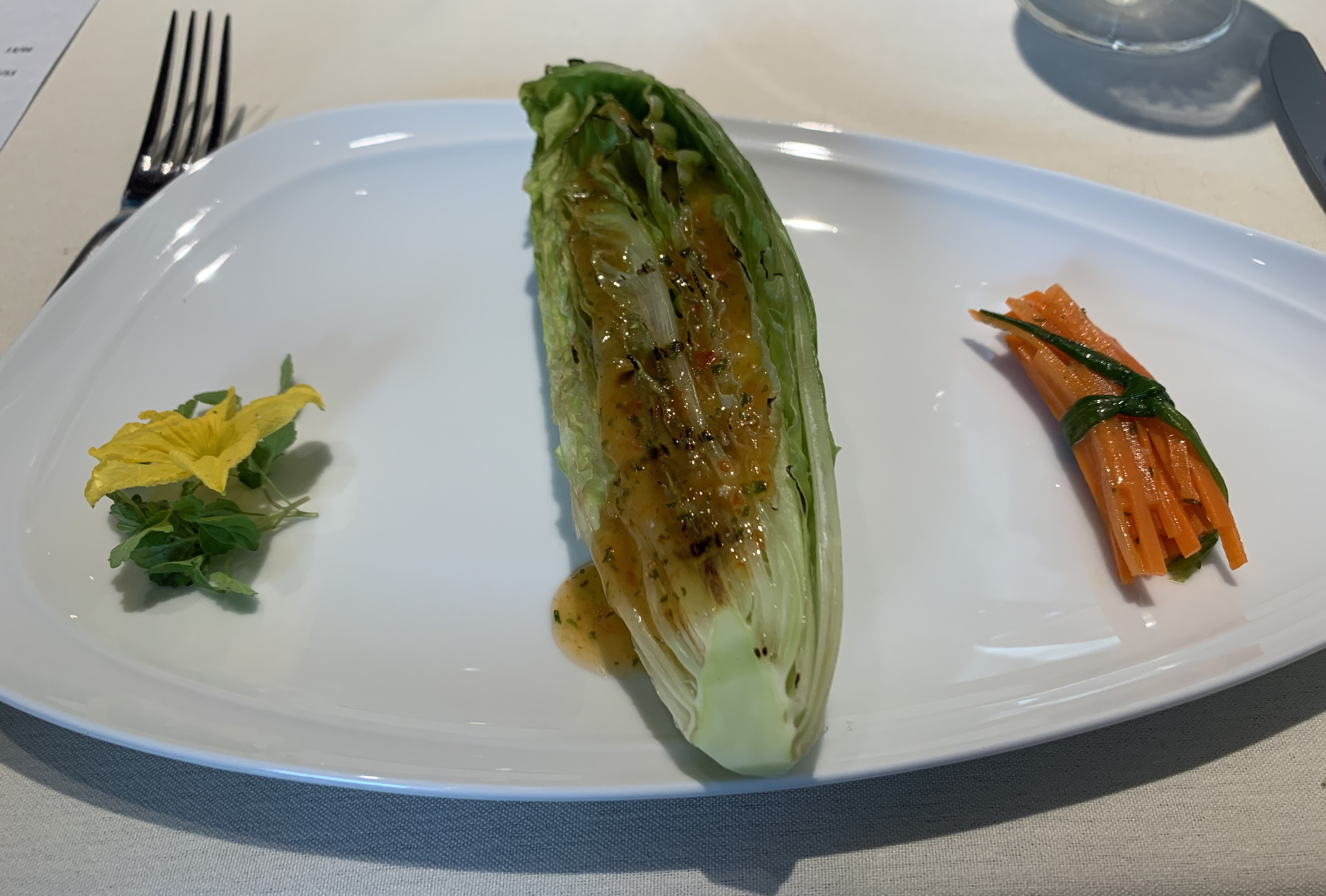 Romaine heart with the leaves still attached that has been cut in half and grilled, with visible grill marks on the lettuce. The lettuce has been dressed with a brown sauce. A bundle of shiny, sauced carrots is next to it, tied off with a wilted green onion. 