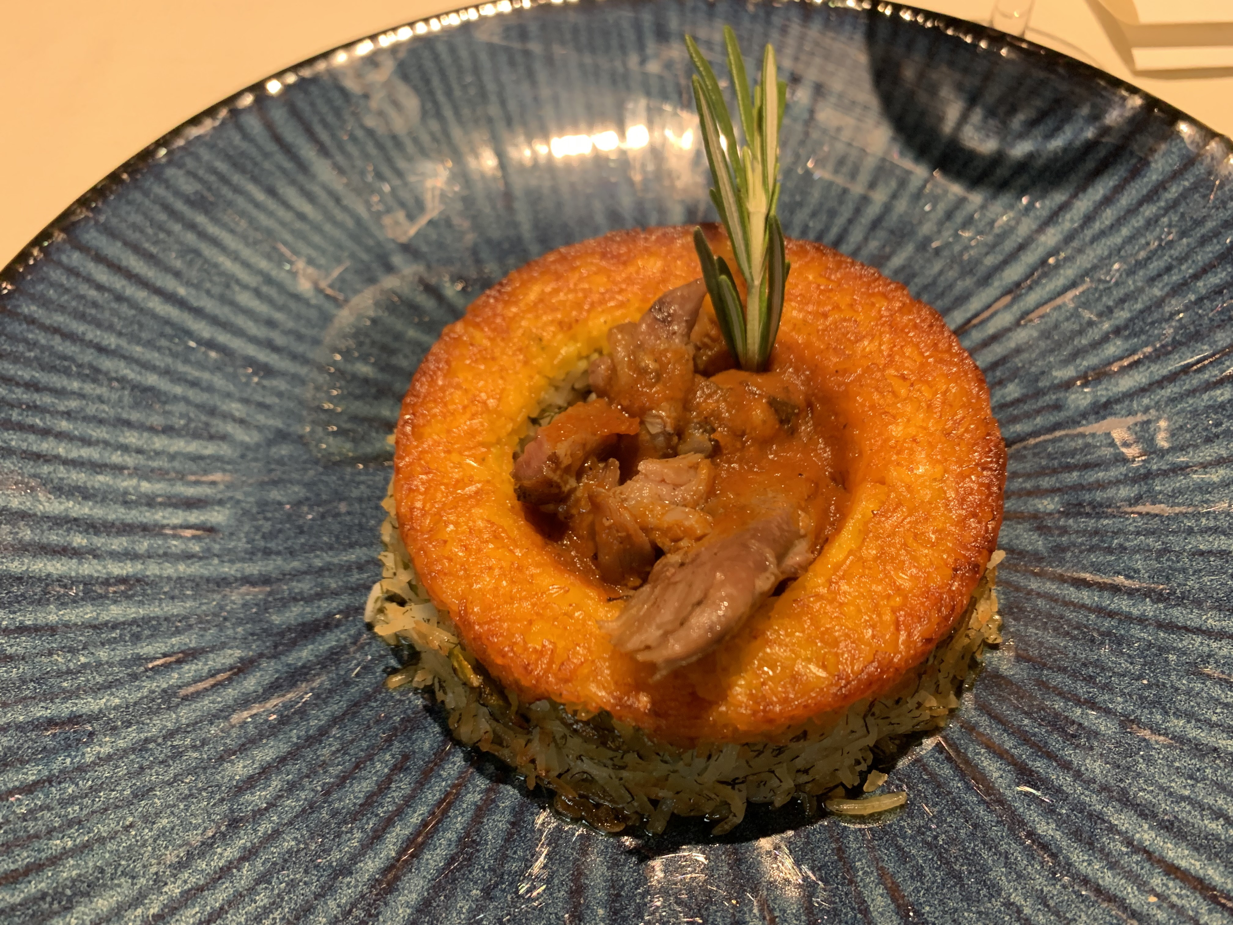 Rice plated in a donut shape, with a sprig of rosemary sticking out. In the middle of the donut is lamb with an orange sauce. The top ring of rice has been cooked to a golden-brown crust.