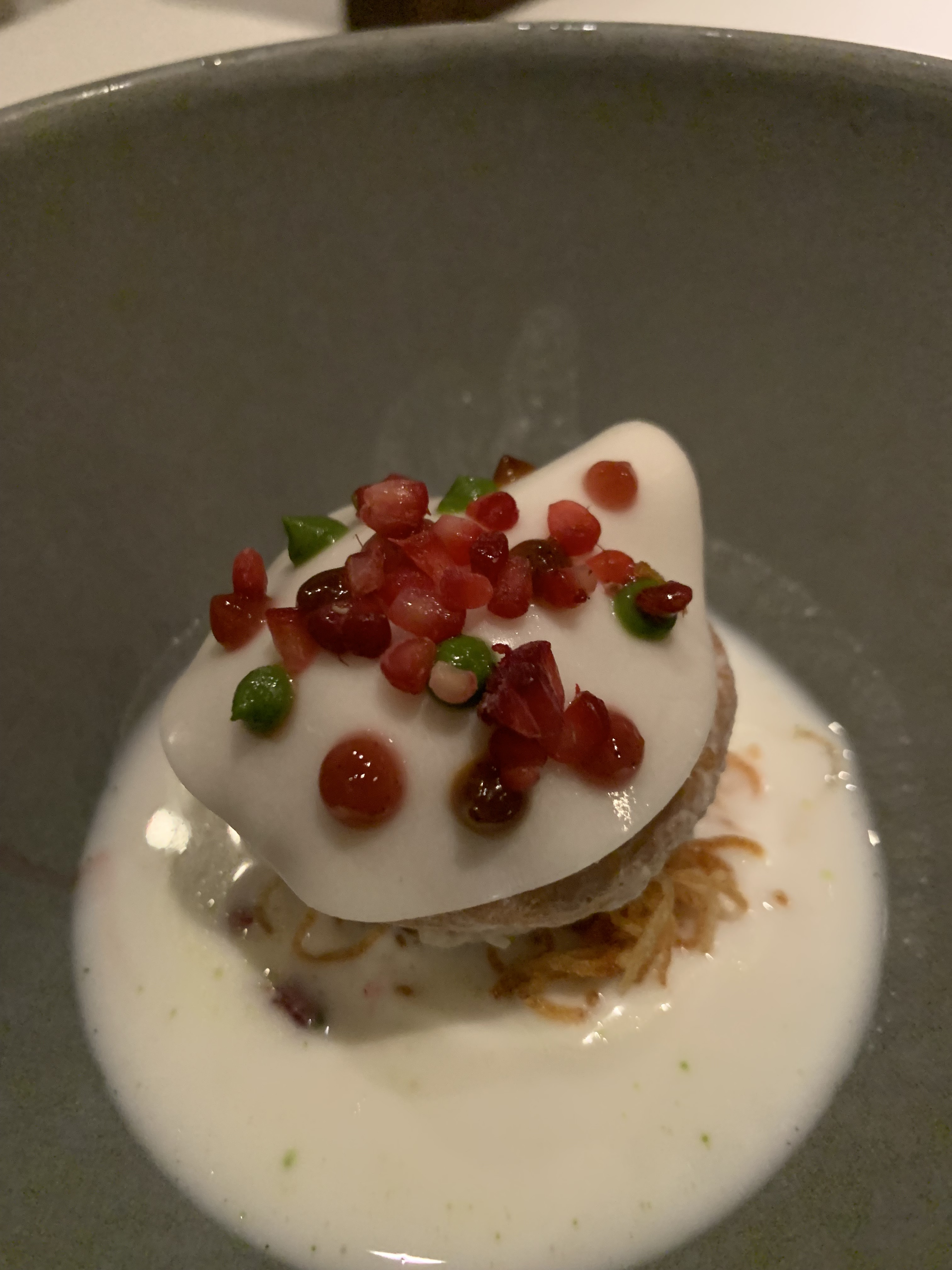 Small dish holding a thin yogurt-based broth, with bits of crispy light-brown tendrils visible below a round pastry topped with a thin, white, shiny gel. Green & red dots of sauce garnish the gel.