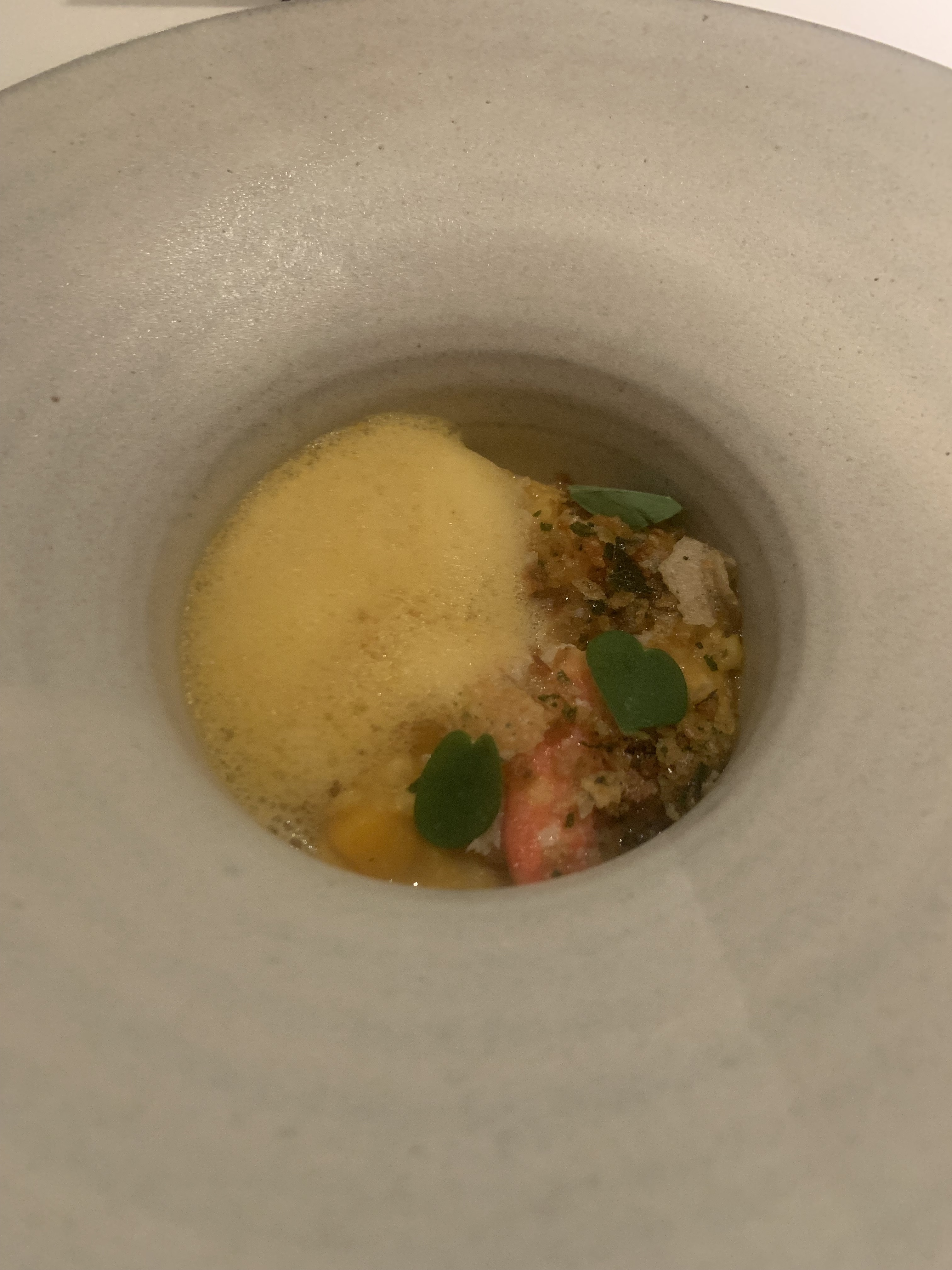 A small bowl with yellow foam covering most of the contents, with the pink & white flesh of crab barely peaking through, and some dark-brown crispy bits mixed in with the crab. A few small, dark-green leaves garnish the crab.