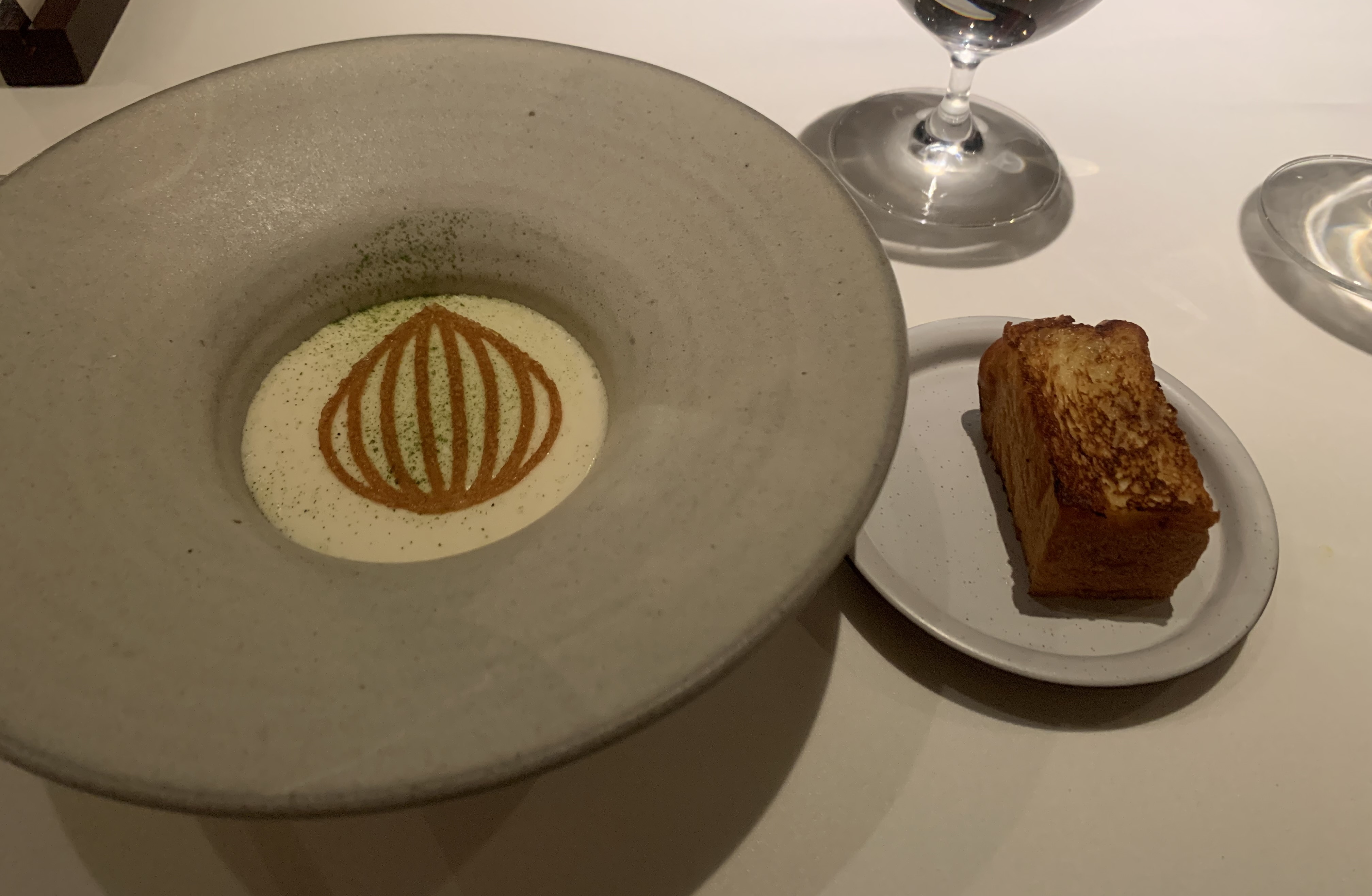 Bowl of white foam with a delicate, dark-brown tuile cookie in the shape of an onion sitting on top, dusted lightly with a green powder. Next to it is a golden-brown toasted hunk of buttery brioche bread.