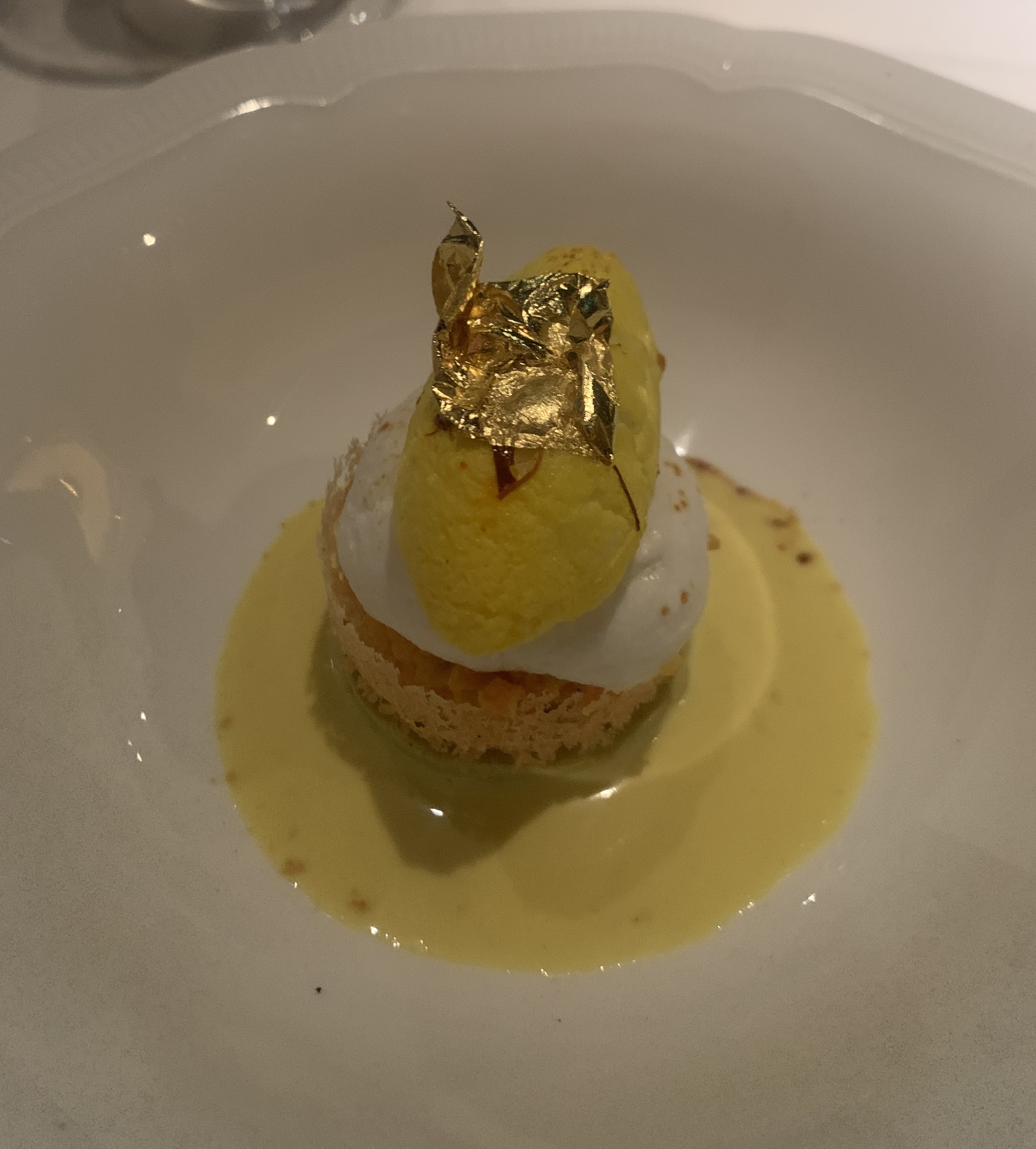 A tower of treats: a delicate shell of pastry on the bottom holding an orange filling, a milk cloud on top of that, and a quenelle of yellow ice cream with golf leaf. Around the shell is a smooth, yellow sauce.