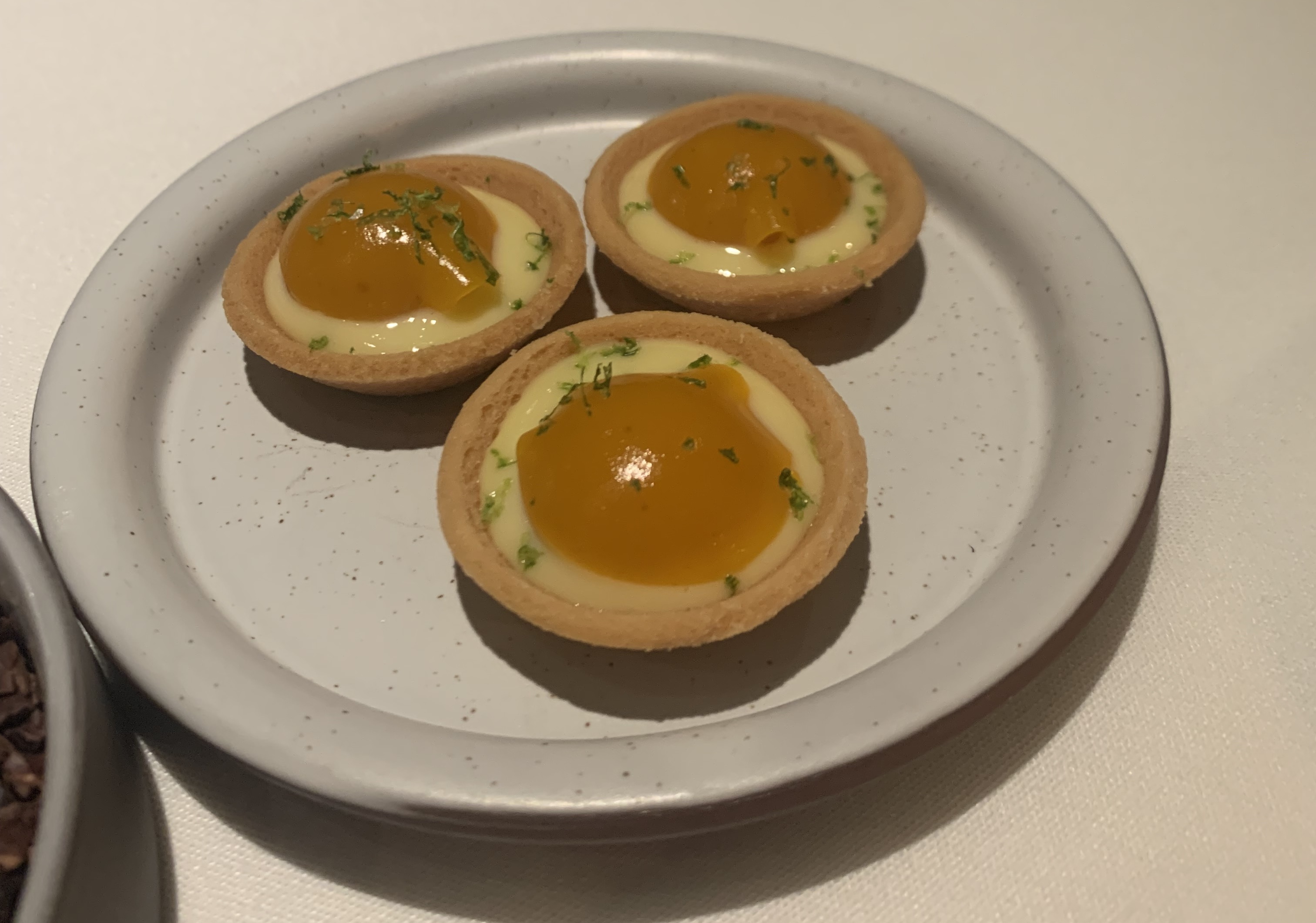 Plate with three round tartlet shells, each filled with a traditional-looking pale-yellow pastry cream. On top of the cream is a smooth, rounded mound of an orange gel. The whole thing looks a bit like an egg cracked into a tart shell.