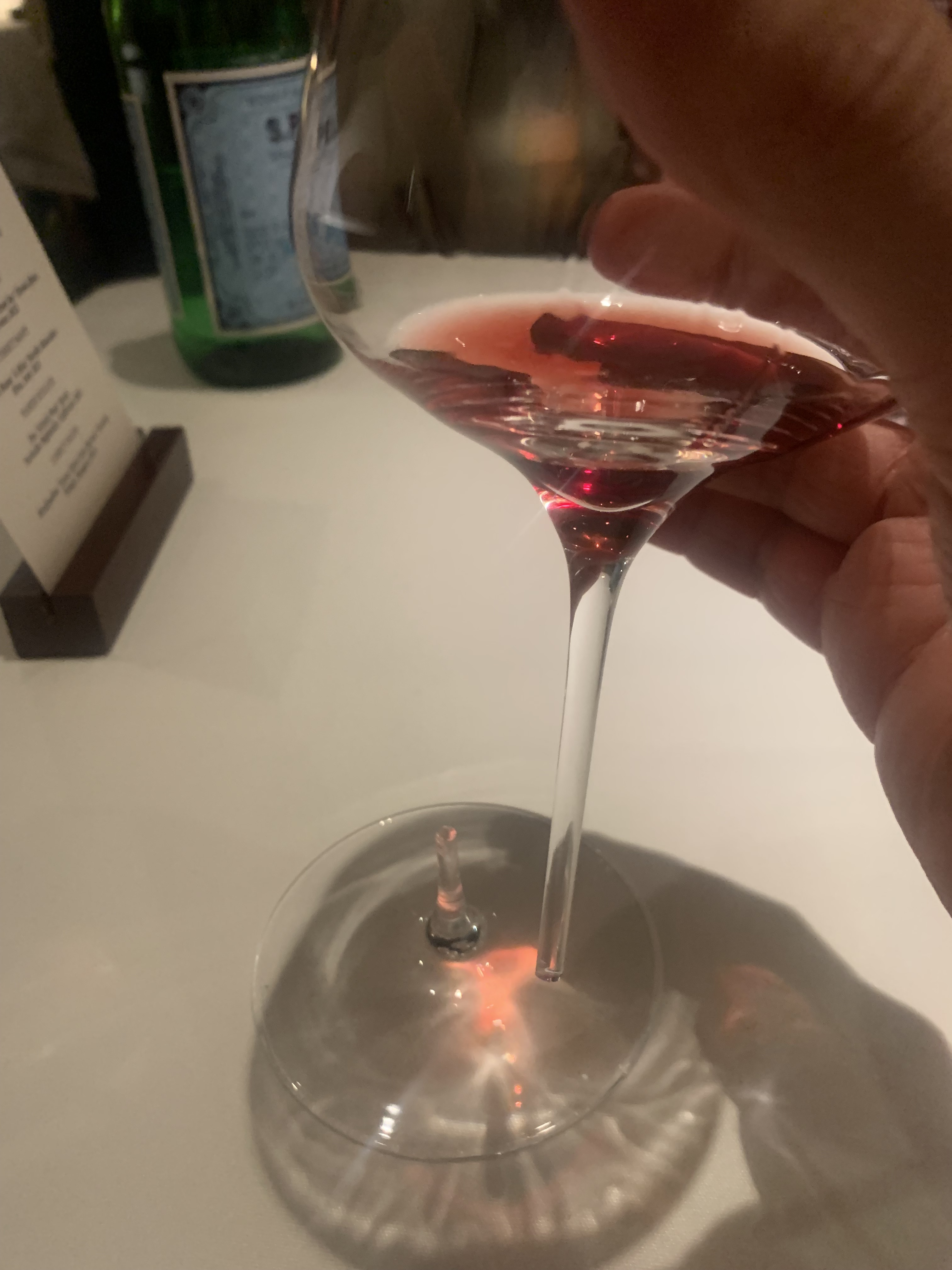 A wine glass with a very thin stem. The stem has broken, and a hand holds the wine glass while the base sits on the table. There is still a little bit of a light red wine in the glass.