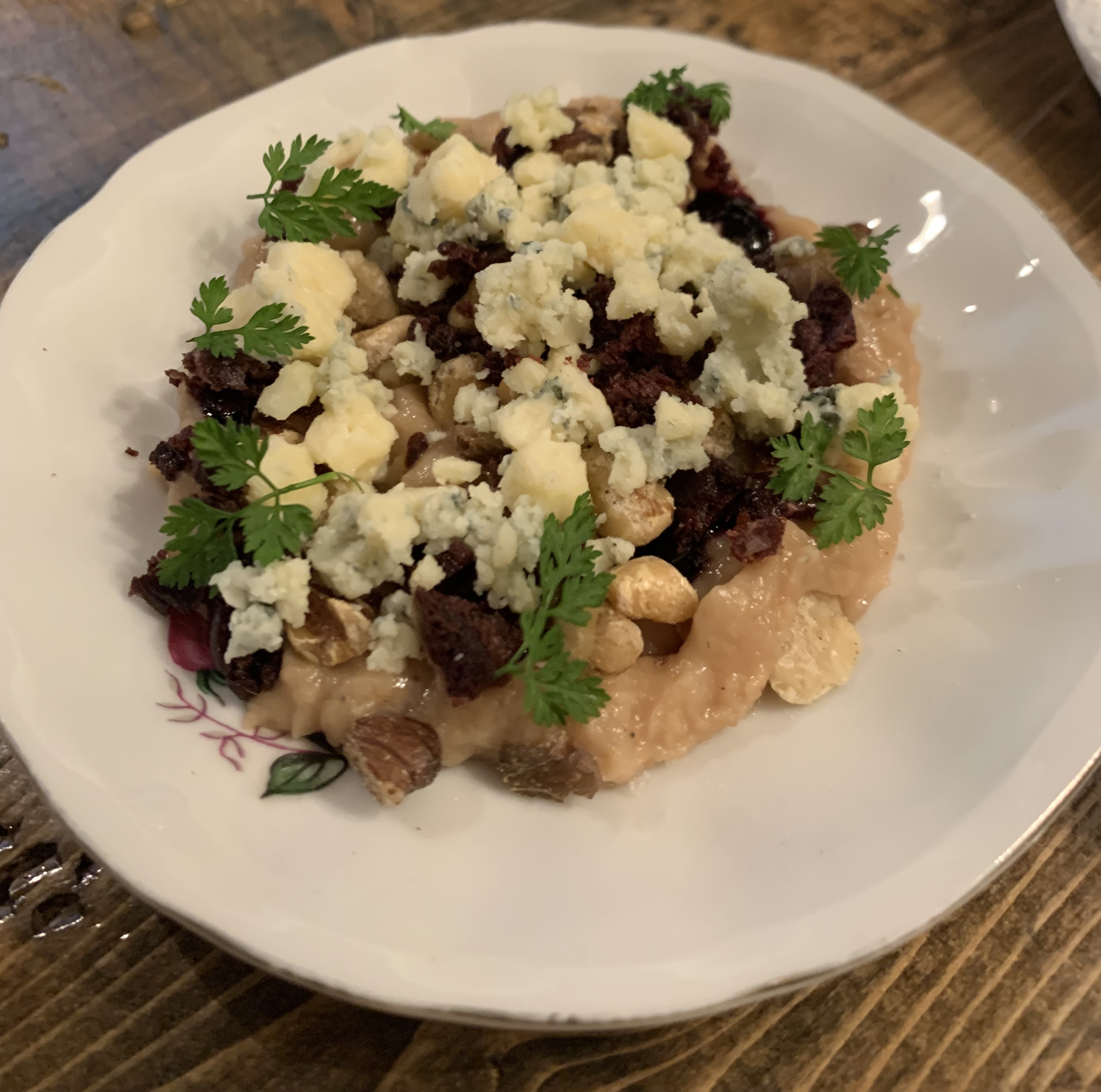 Mashed white bean dip base, with blue cheese crumbles, tiny sprigs of herbs, little bits of walnuts, and dried bits of raspberry on top of it.