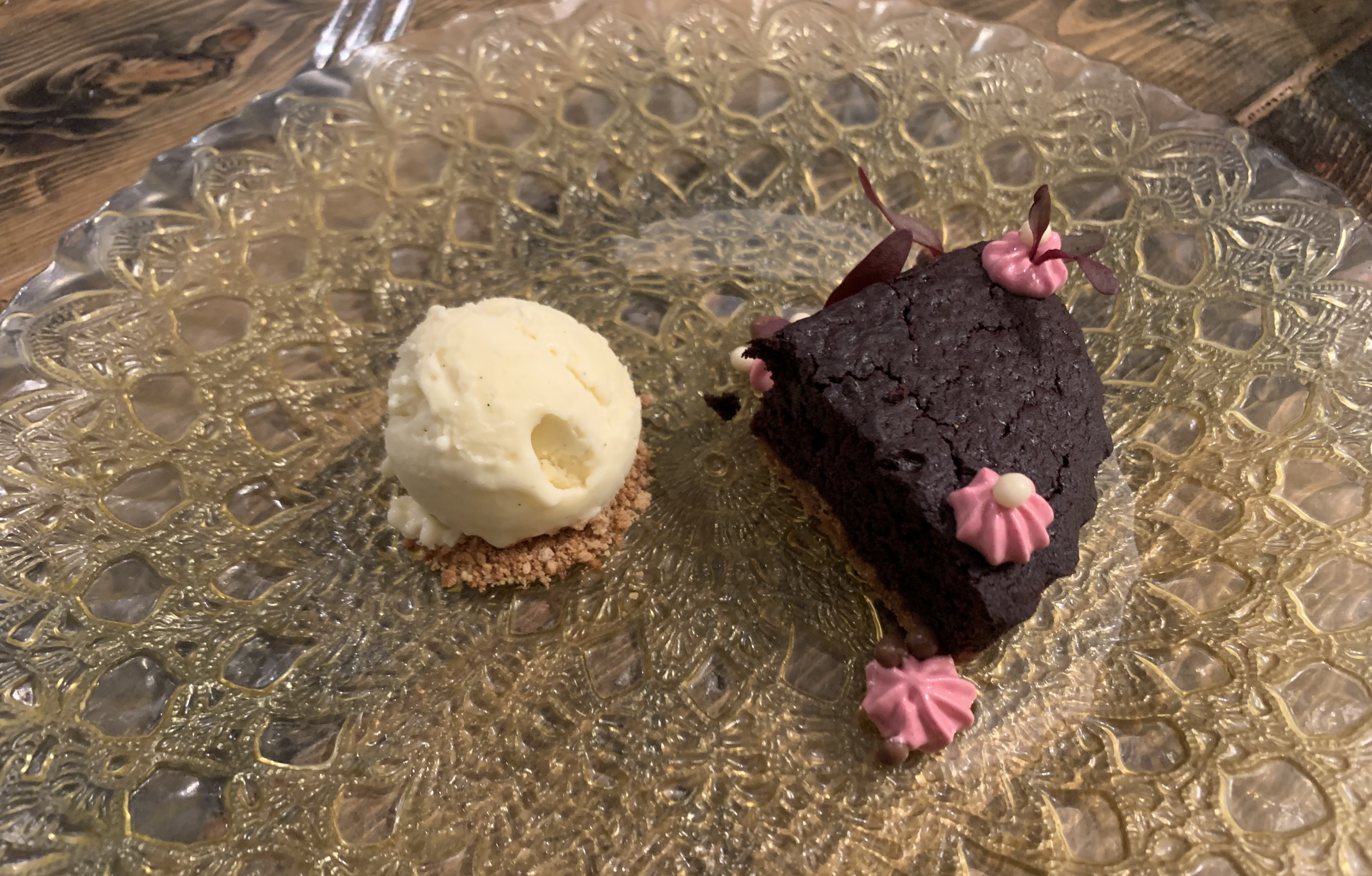 Large translucent plate with golden scrollwork. On the left is a scoop of perfectly white ice cream atop a bed of crushed graham cracker. On the right is a thick slice of brownie pie, dark black, and topped with a few dots of pink frosting made to look like flowers.