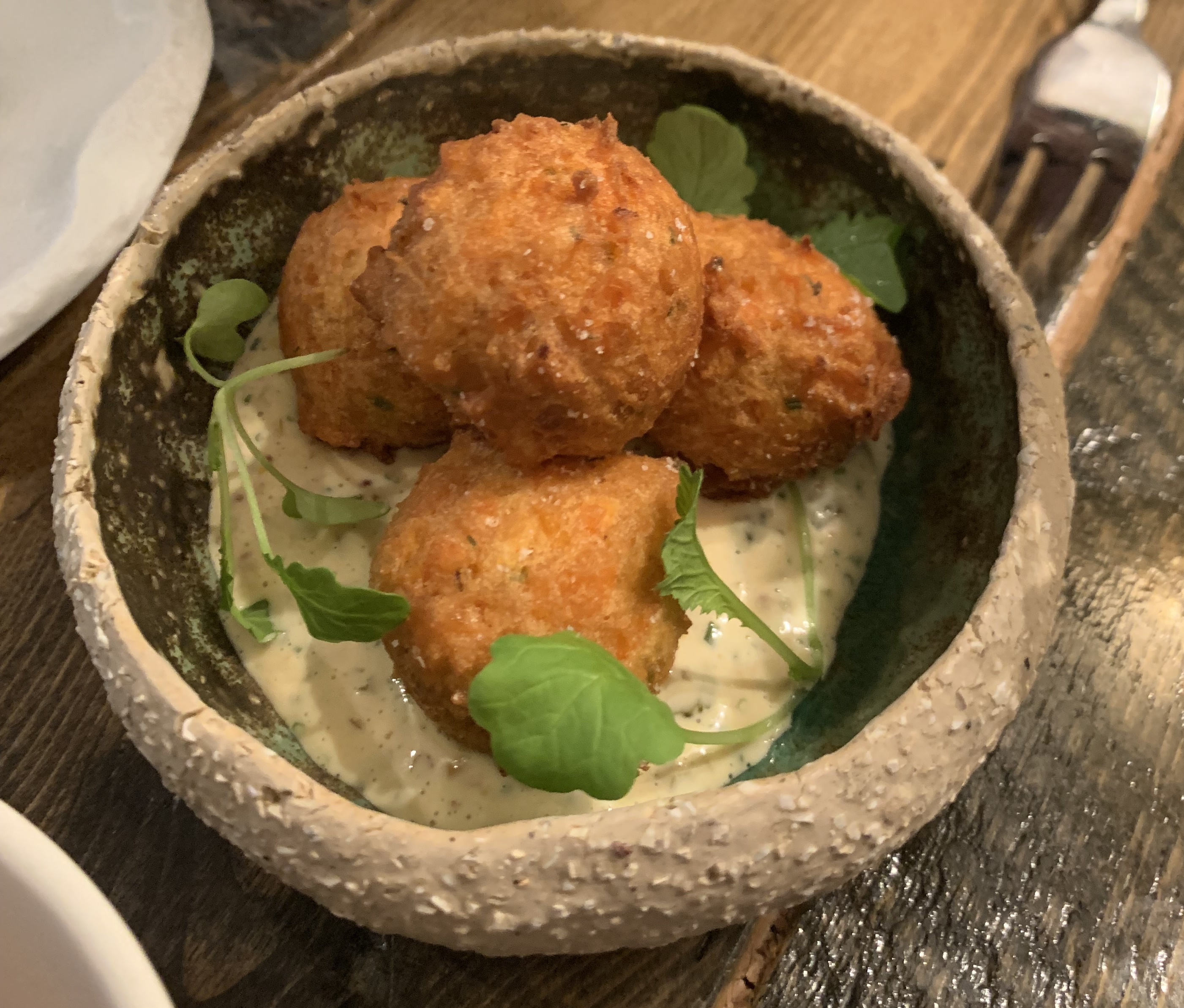 A bowl of four fried balls, crispy and golden. Three balls sit in a pale sauce with flecks of green in it; the fourth ball sits on top of the other three, forming a pyramid. The dish is garnished with three sprigs of delicate-looking green herbs with wide leaves.