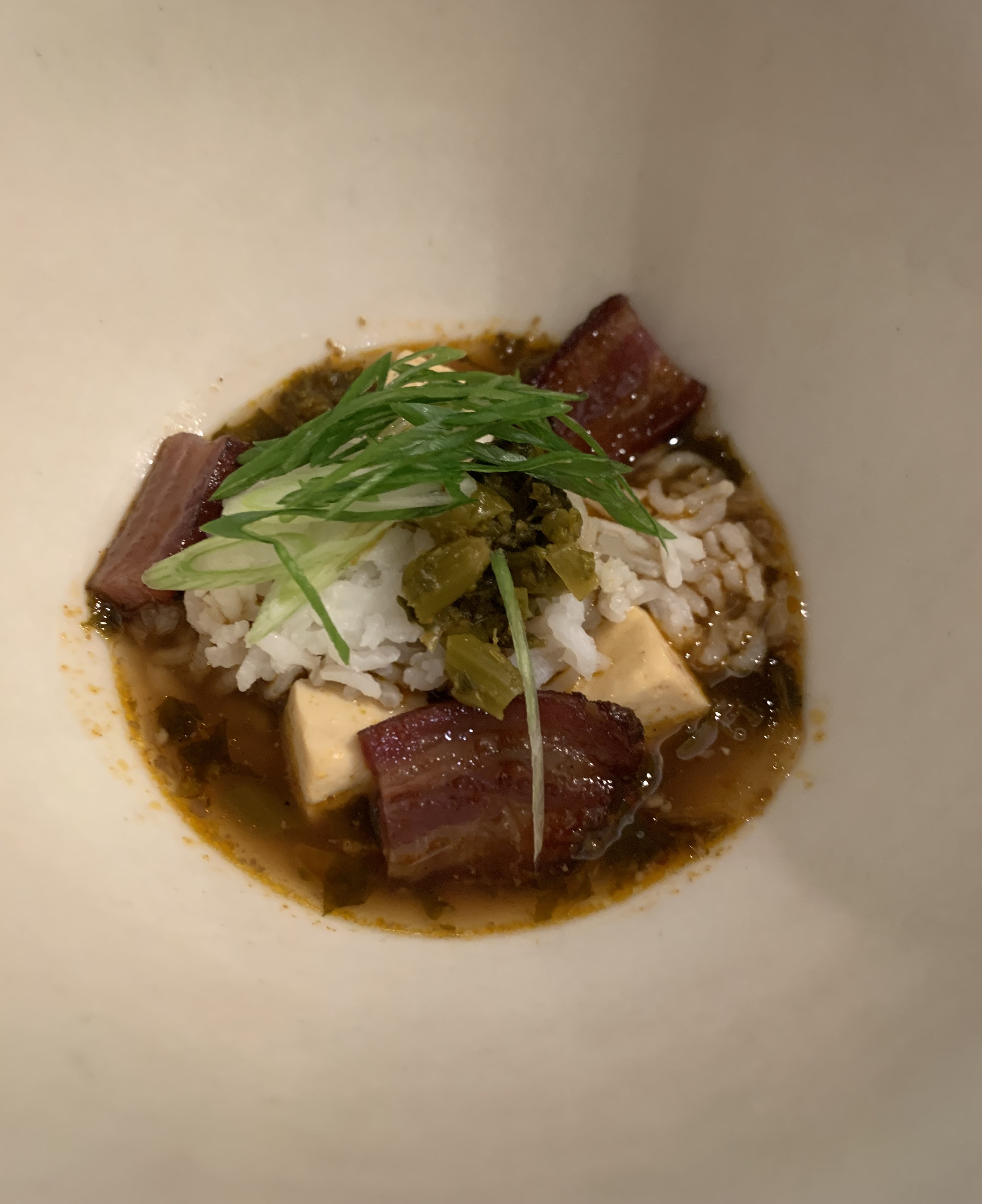 Rice topped with big, thick chunks of bacon. The bacon has charred edges. There's fresh, super-thinly-sliced green onion on top of the rice, along with a green pickled vegetable. Some pale yellow cubes of tofu are visible beneath the rice. There is a shallow pool of dark-gold broth underneath everything.
