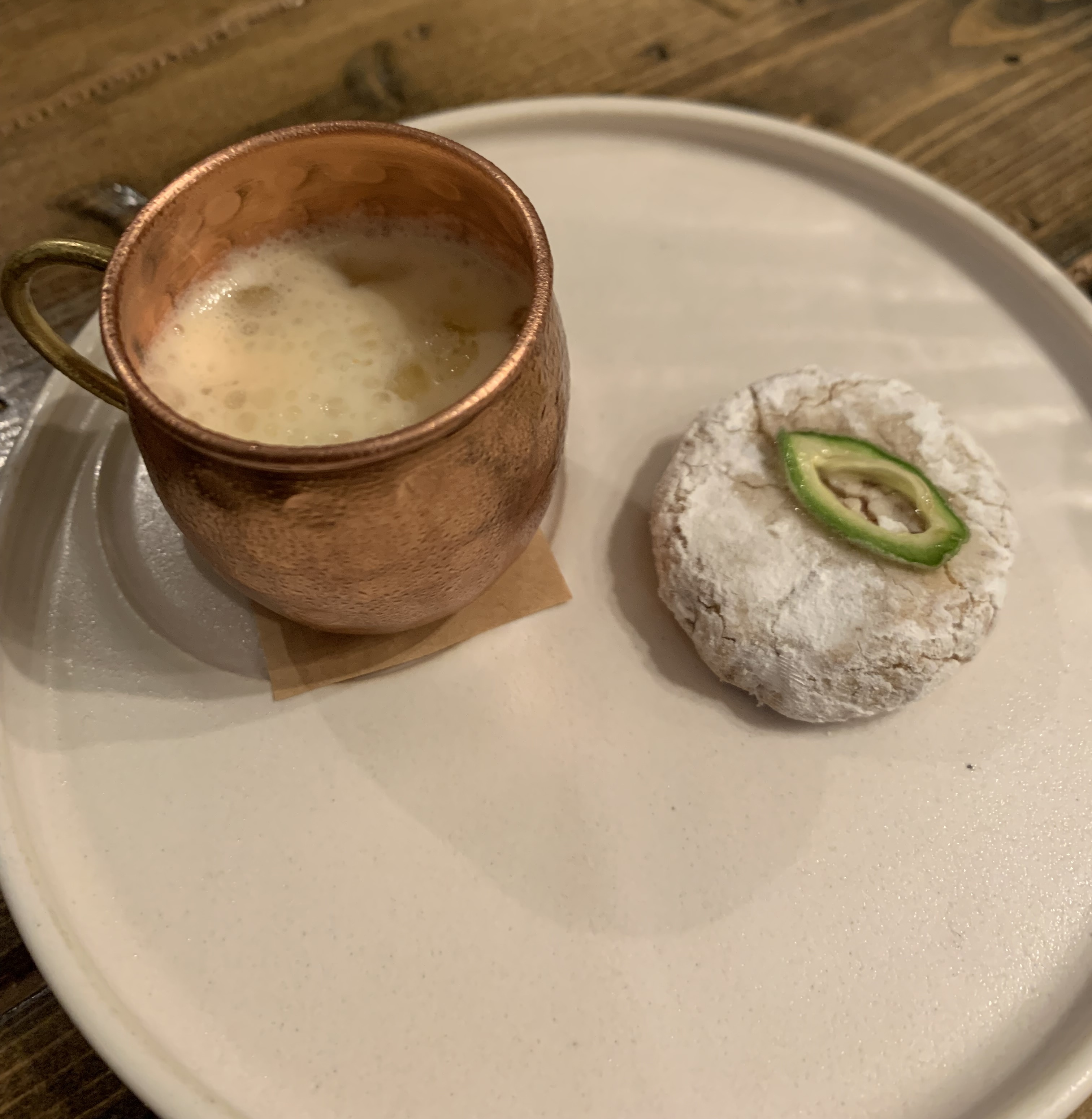 A plate with a tiny, hammered-copper cup with an orange-tinted milk and tiny ice cubes. Next to it is a cookie covered in white powder, with a single slice of a green almond on top.