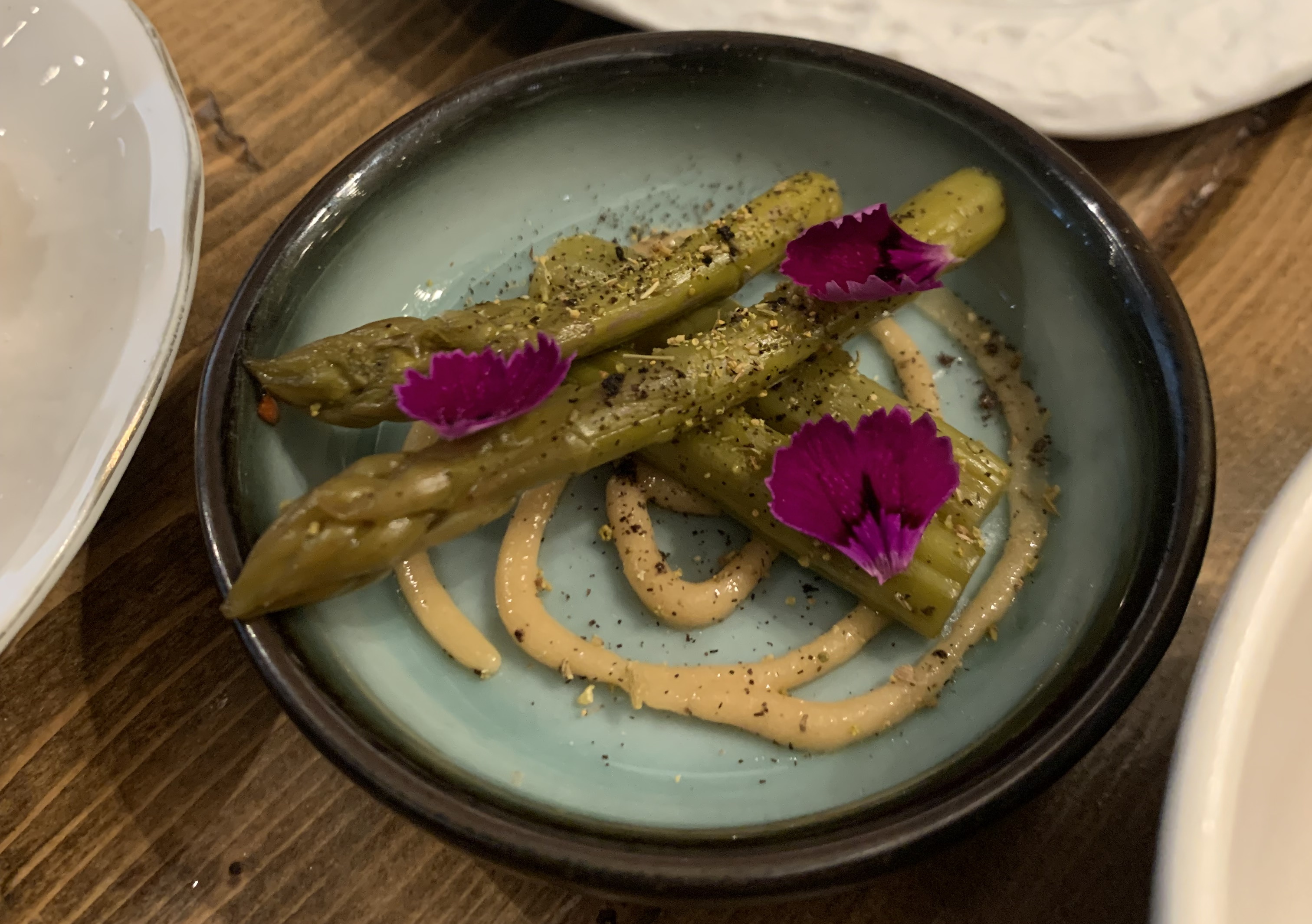Four thin spears of asparagus arranged in two layers, with the tips perpendicular to the bodies of the spears. There's a lot of seasoning on the spears, and underneath everything is a drizzle of lightly-colored sauce, holding its shape. The dish is garnished with purple flower pedals.
