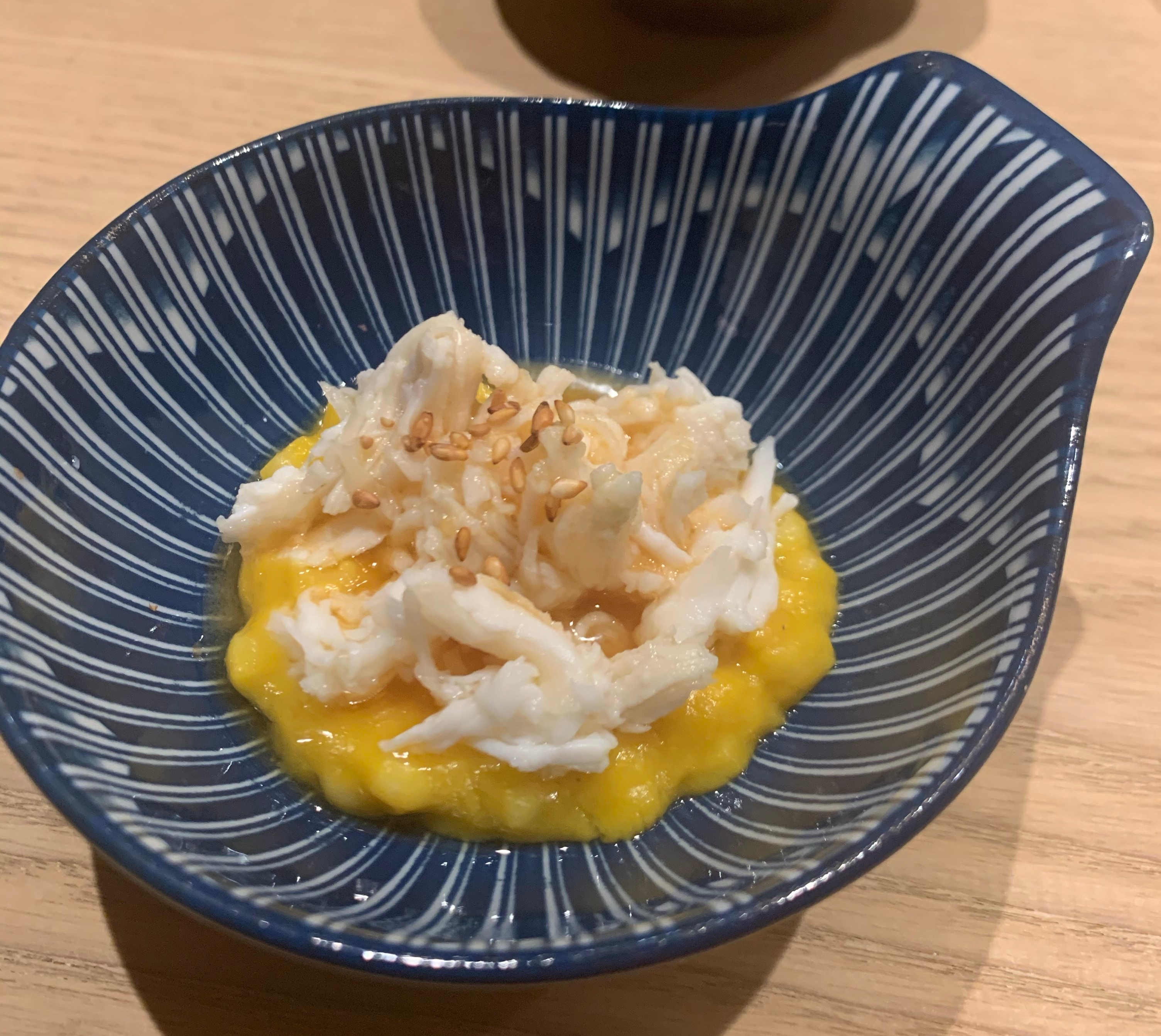 A bowl (shaped like a scallop shell) with a mound of corn-yellow puree, topped with large chunks of white crab meat. There are a few toasted sesame seeds for garnish.