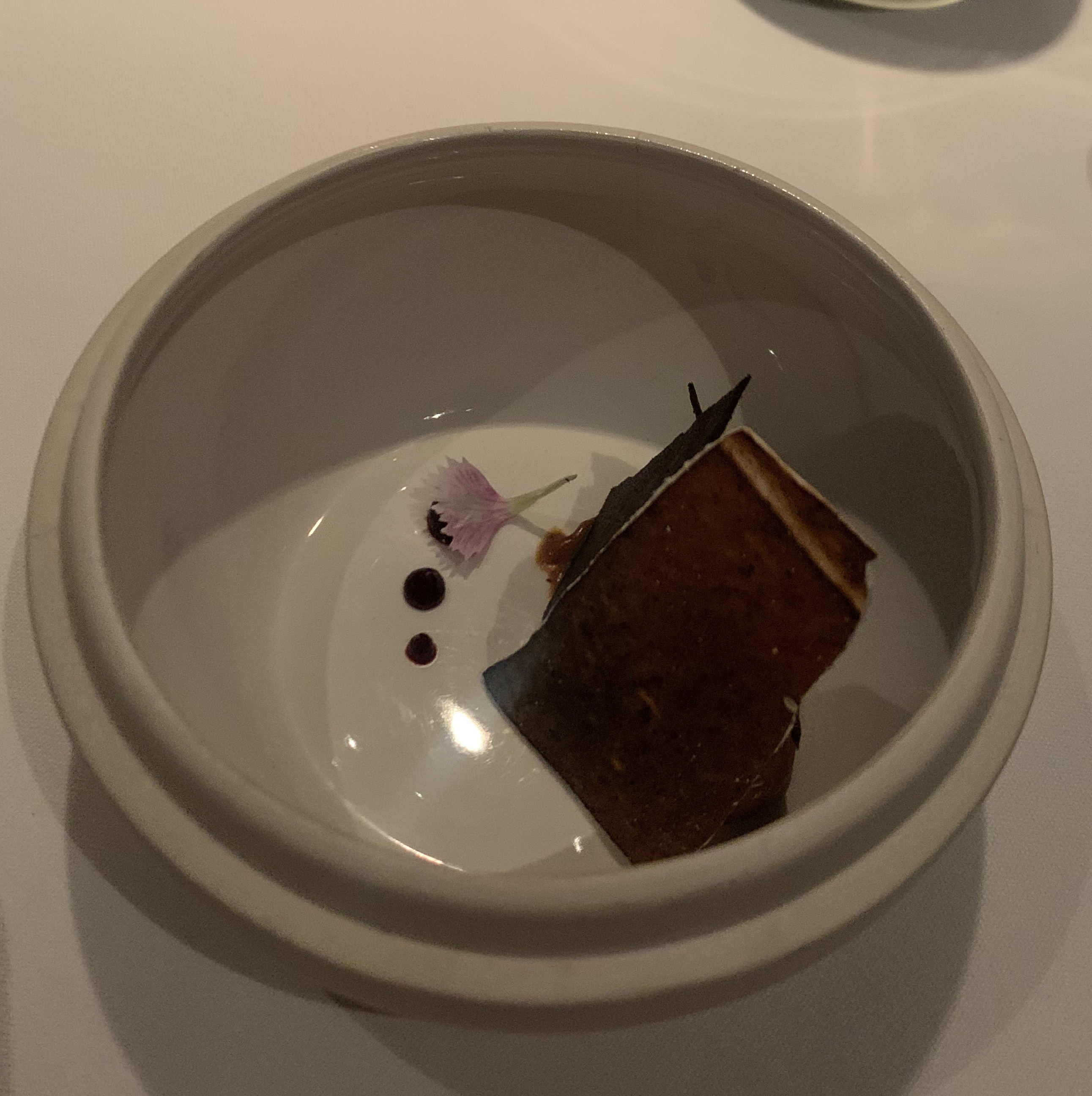Off-white bowl containing some sheets of dark brown merigune standing up, hiding something. Next to them are three dots of chocolate sauce, with the top dot being larger and the two below it decreasing in size. There is one pink flower garnishing the dish, obscuring the top chocolate sauce dot.