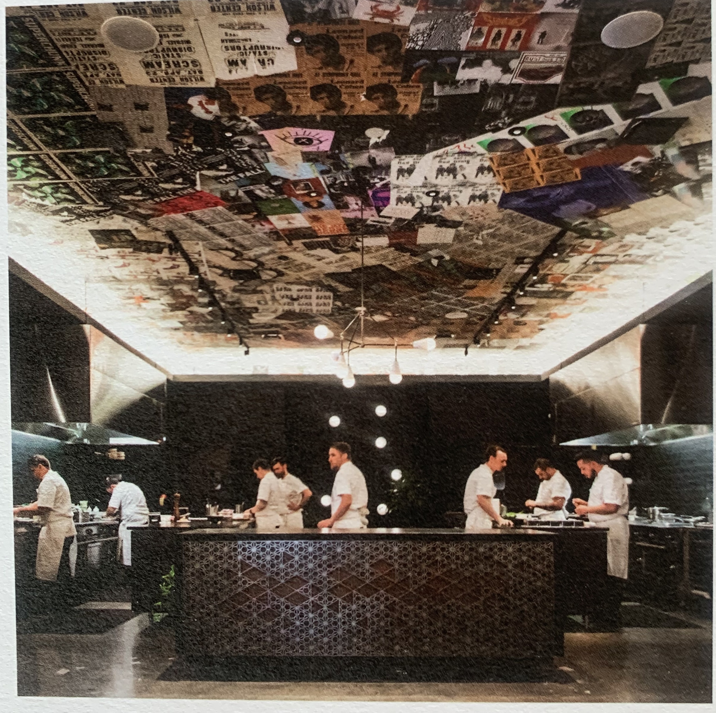 Photo of a printed image of the kitchen. It's an open kitchen, with an island at the front and cooking stations along either wall. The ceiling is well-lit around its perimeter, illuminating a ton of posters for bands & performances that adorn it. The posters are arranged somewhat randomly as a collage, with blocks of the same poster in some spots. Eight chefs in white jackets work diligently in the kitchen.