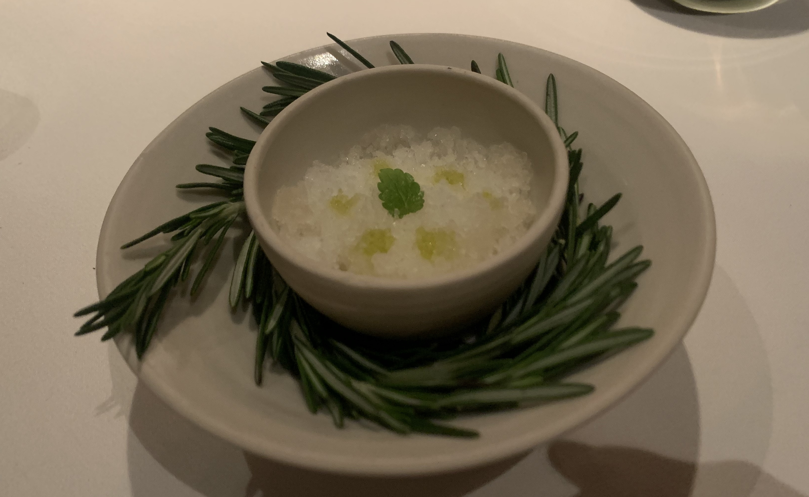 Saucer with a bowl of shaved ice on top of it. The bowl of shaved ice is embraced by sprigs of rosemary wound around it like a nest. The shaved ice itself is clear ice with a few dollops of yellow, and one single mint leaf in the middle.