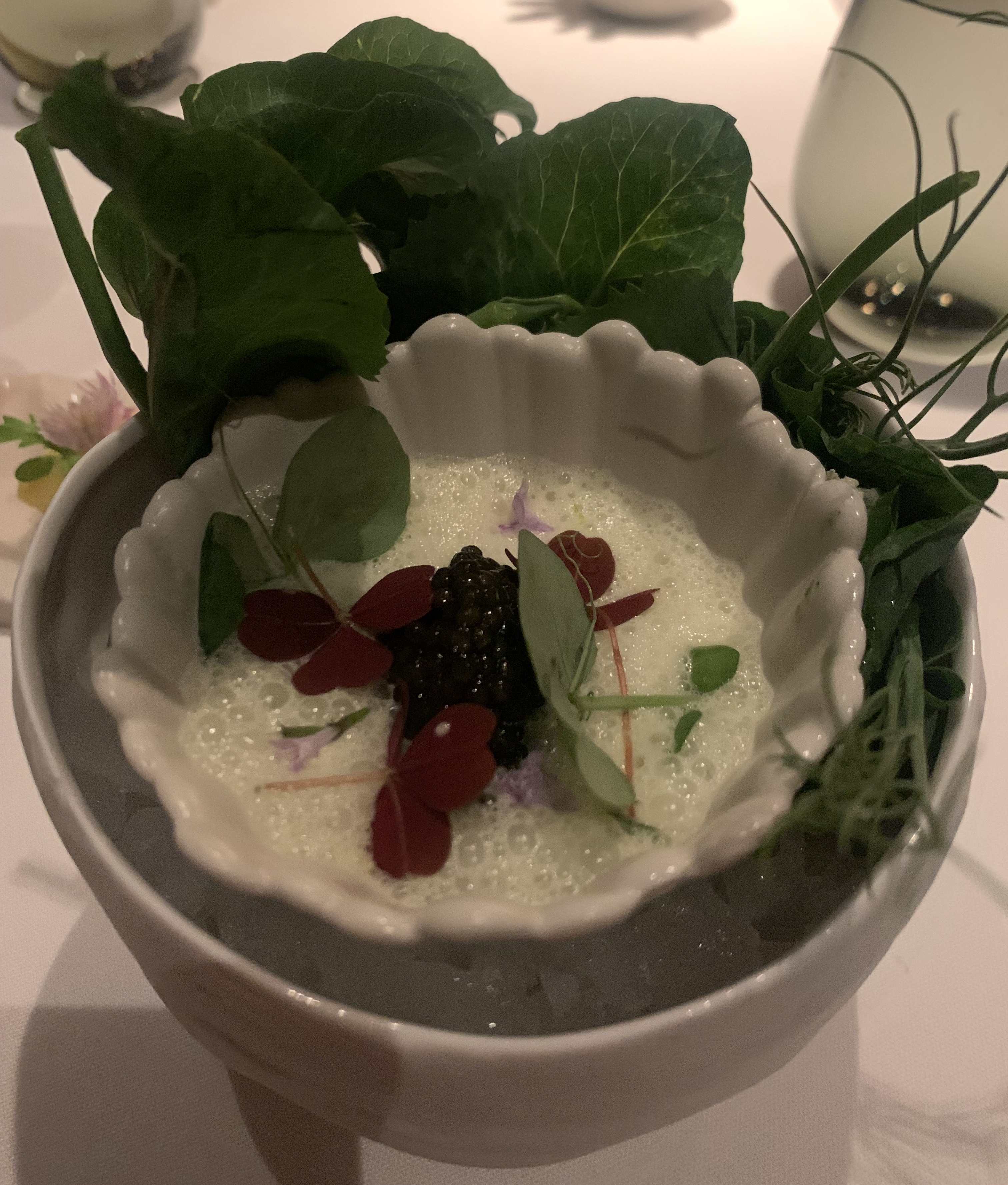 Larger bowl of ice containing a smaller bowl with a mound of caviar atop a lot of white foam. There are tons of greens shoved between the two bowls, and garnishing the foam are pea flowers, greens, and a couple red leaves.