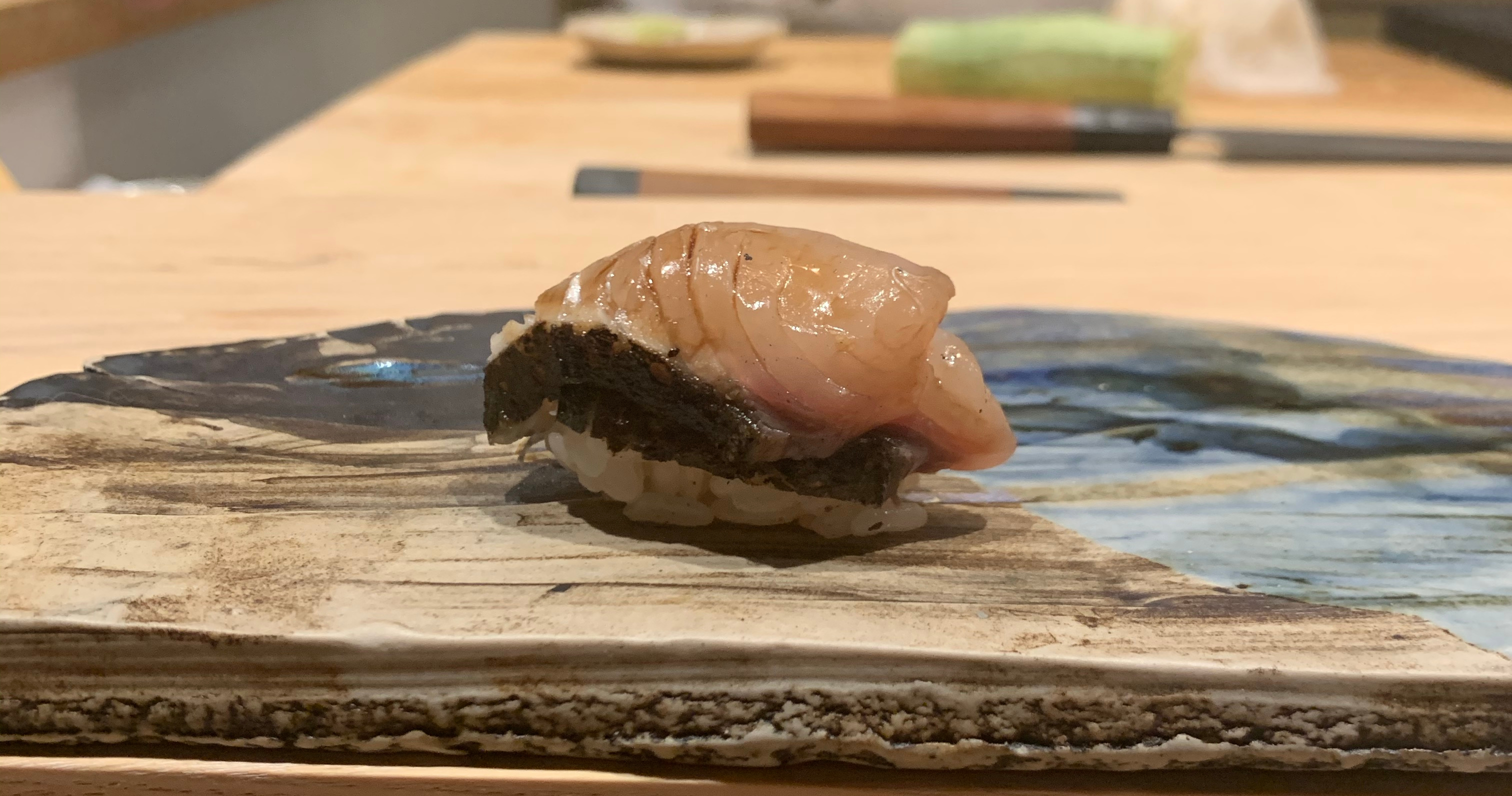 One piece of nigiri sushi with a piece of fish on top. The fish has a band of skin along the bottom. The top of the fish is pink and has been brushed with a soy sauce glaze.