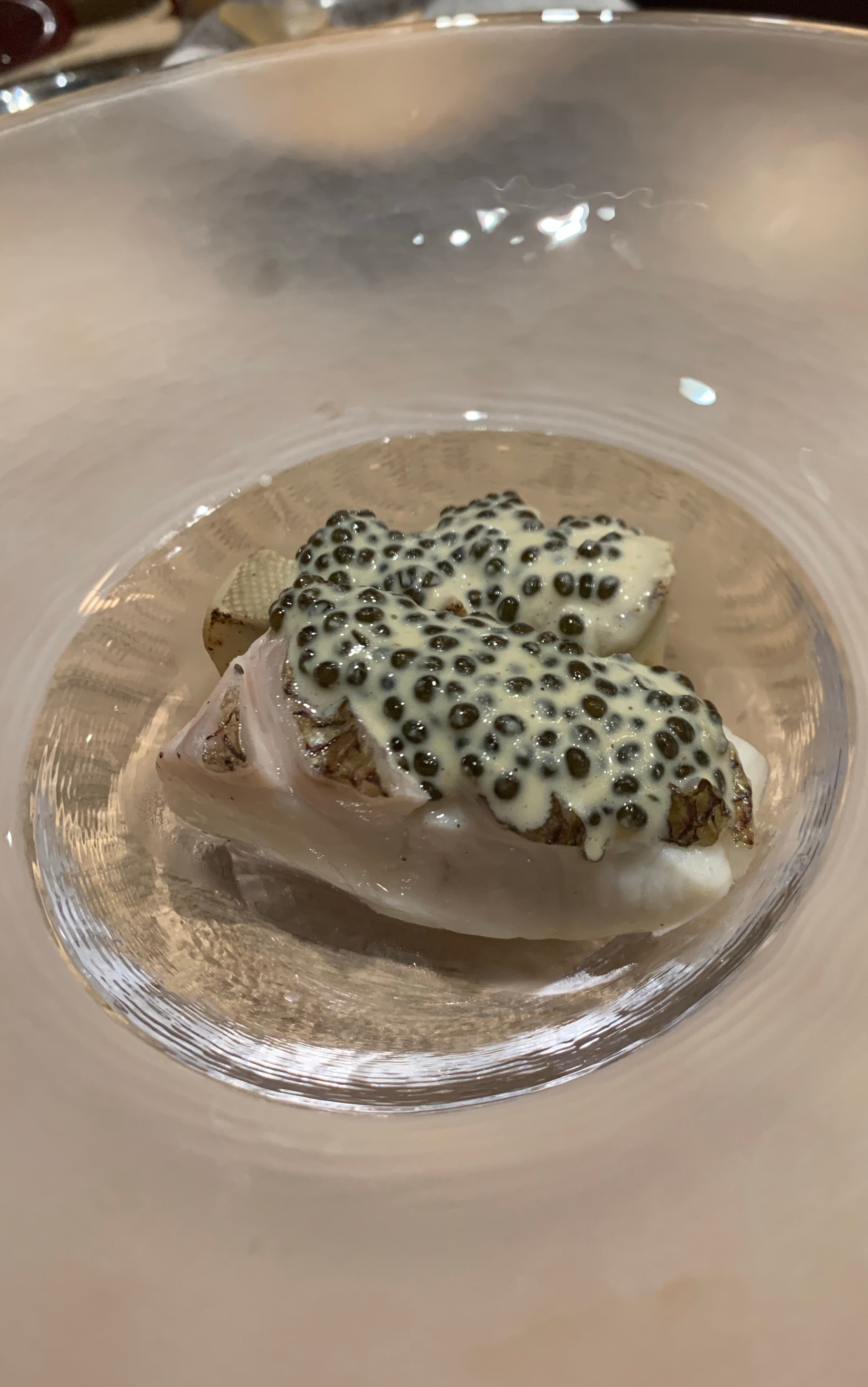 Large plate with a small depression for the food. There is a piece of cooked white fish with a little colour from the puffed scales where they have been left on, topped with caviar and a white sauce.