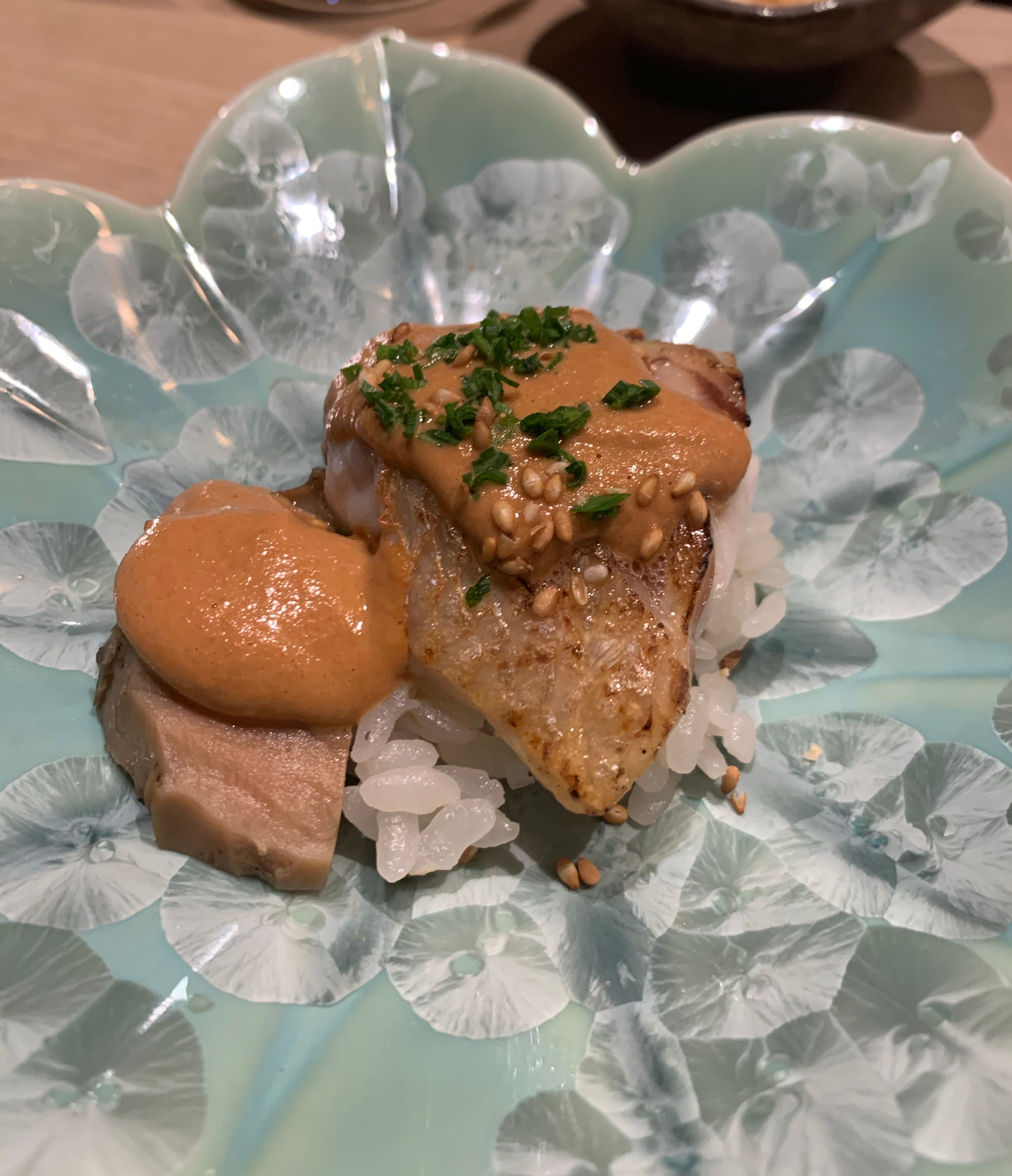 A plate with a light-brown cooked seaperch filet resting on top of a bed of rice. Next to it is a similarly-coloured slice of abalone. Both pieces of seafood have a dollop of orange sauce on top.