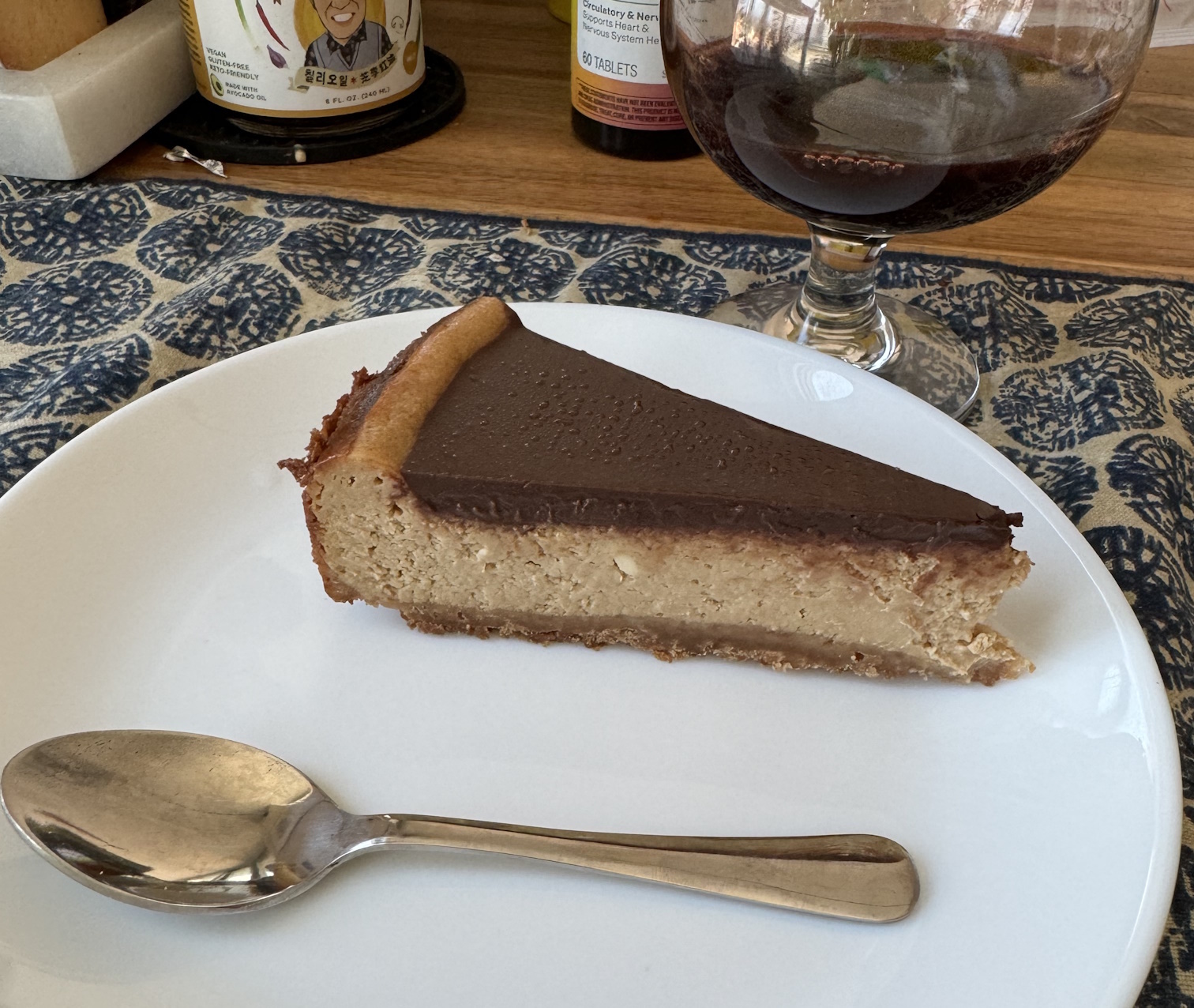 A slice of light brown cheesecake, with a dark brown crust underneath it, and a thick layer of chocolate ganache on top. It's towards the top of a plate, with a shiny steel spoon below it. There is a wide, round-bottomed wine glass behind it filled with dark red port.