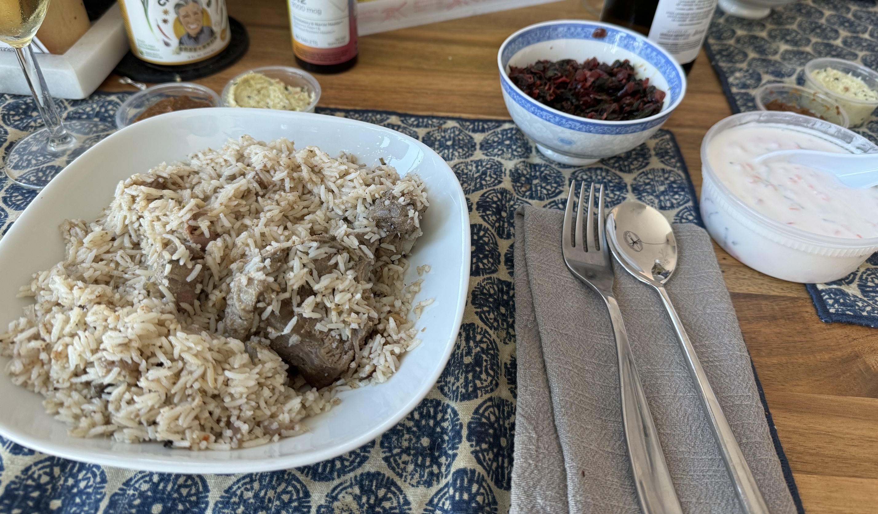 A plate of long-grain rice, with some large chunks of grilled goat barely visible below the surface. Off to the side is a smaller bowl filled with stir-fried greens. There is a large bowl of yogurt with orange carrot flecks in it, a container of a dark reddish-brown paste, and a container of a pale off-white paste.