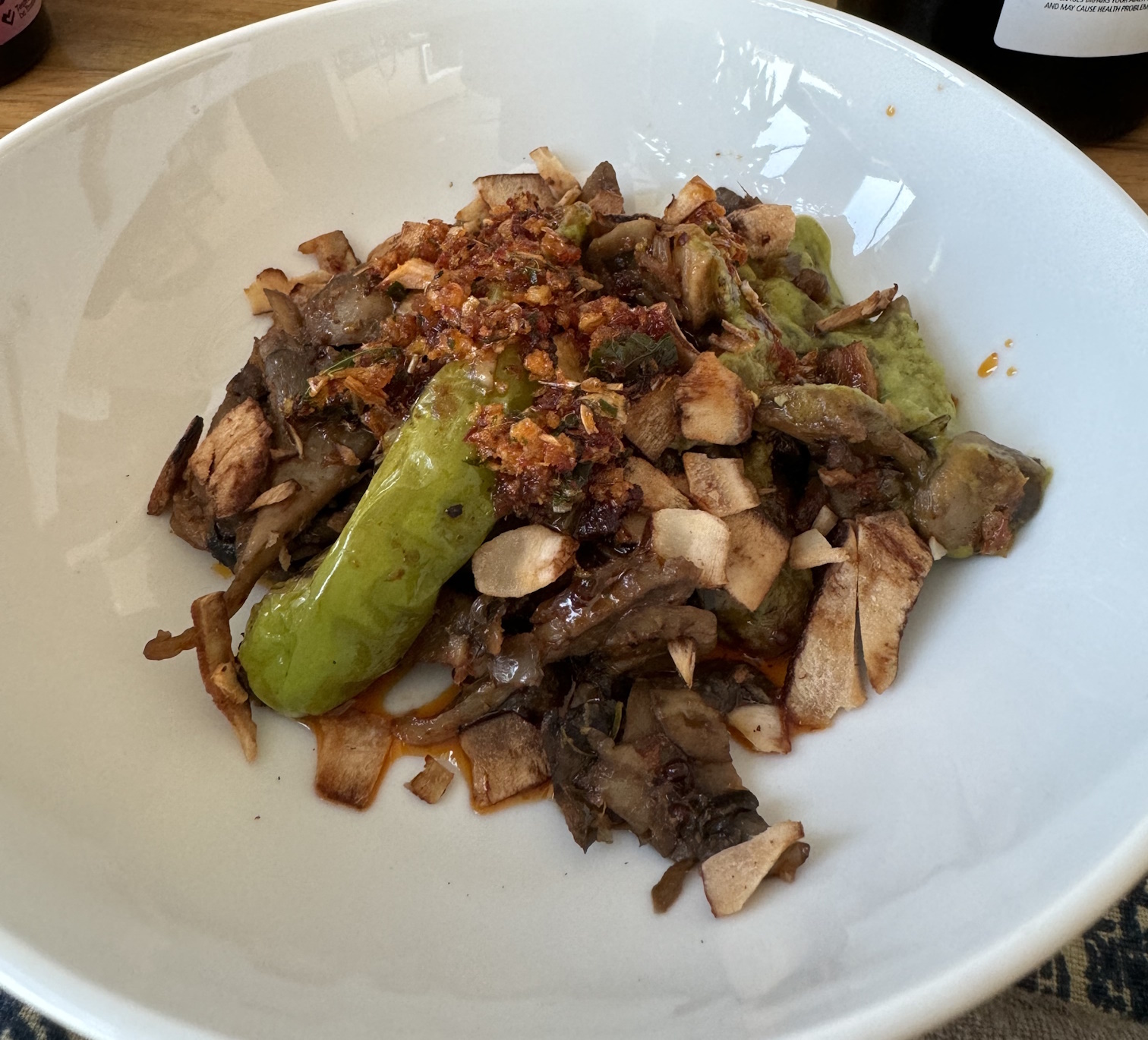 Plate of stir-fried mushroom slices. Beneath is a thick, vibrant green sauce. The dish is topped with chili crisp, thick coconut chips, and a few long shishito peppers blistered black.