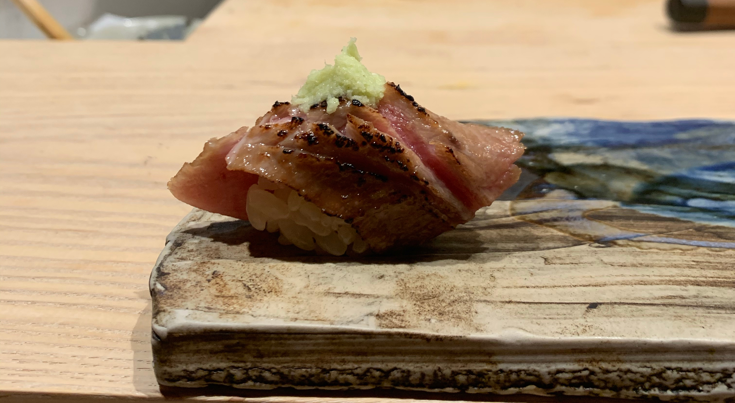 A single piece of nigiri sushi with a seared piece of fish on top. The cooked part is brown, but you can see the red uncooked flesh in some places. It is lightly charred. There is a pile of pale green wasabi on top of the fish.