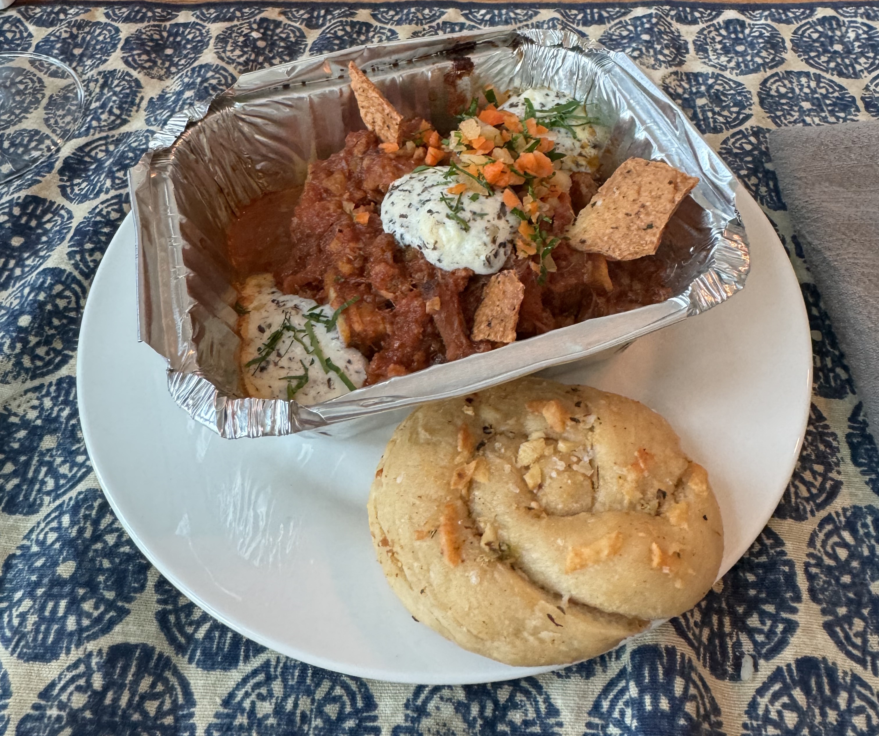 A plate with a metal oven-safe takeout dish and a knot of bread. The tray has several globs of baked cheese dotting a red meat sauce. There are herbs and a fresh veggie garnish. The garlic knot has some crispy parmasean baked on top.