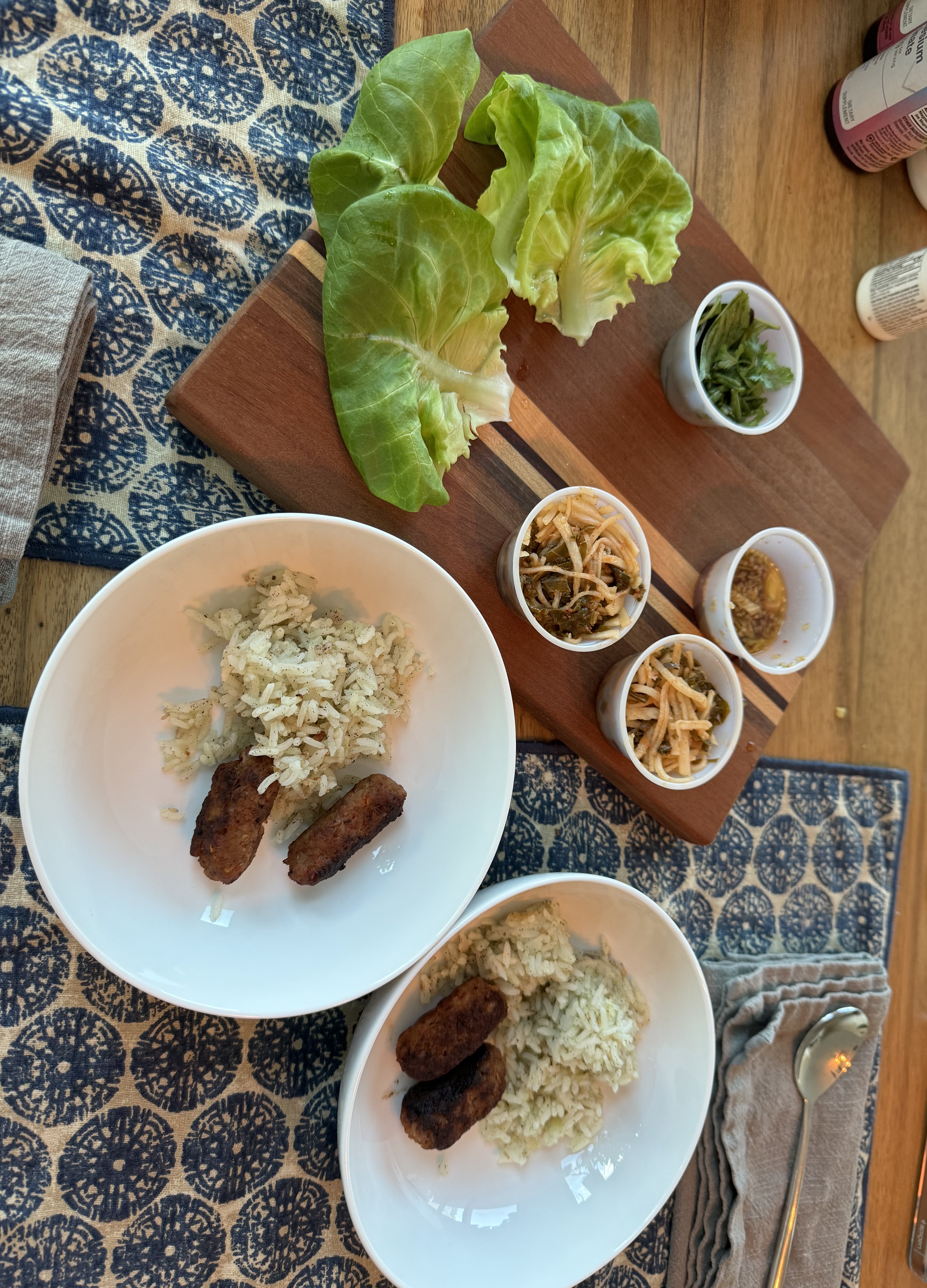 Two plates with a small heap of seasoned rice next to two stout, well-charred case-less sausages. Next to the plates is a cutting board with big, vibrant green whole lettuce leaves and cups of herbs, kimchi, and other toppings.