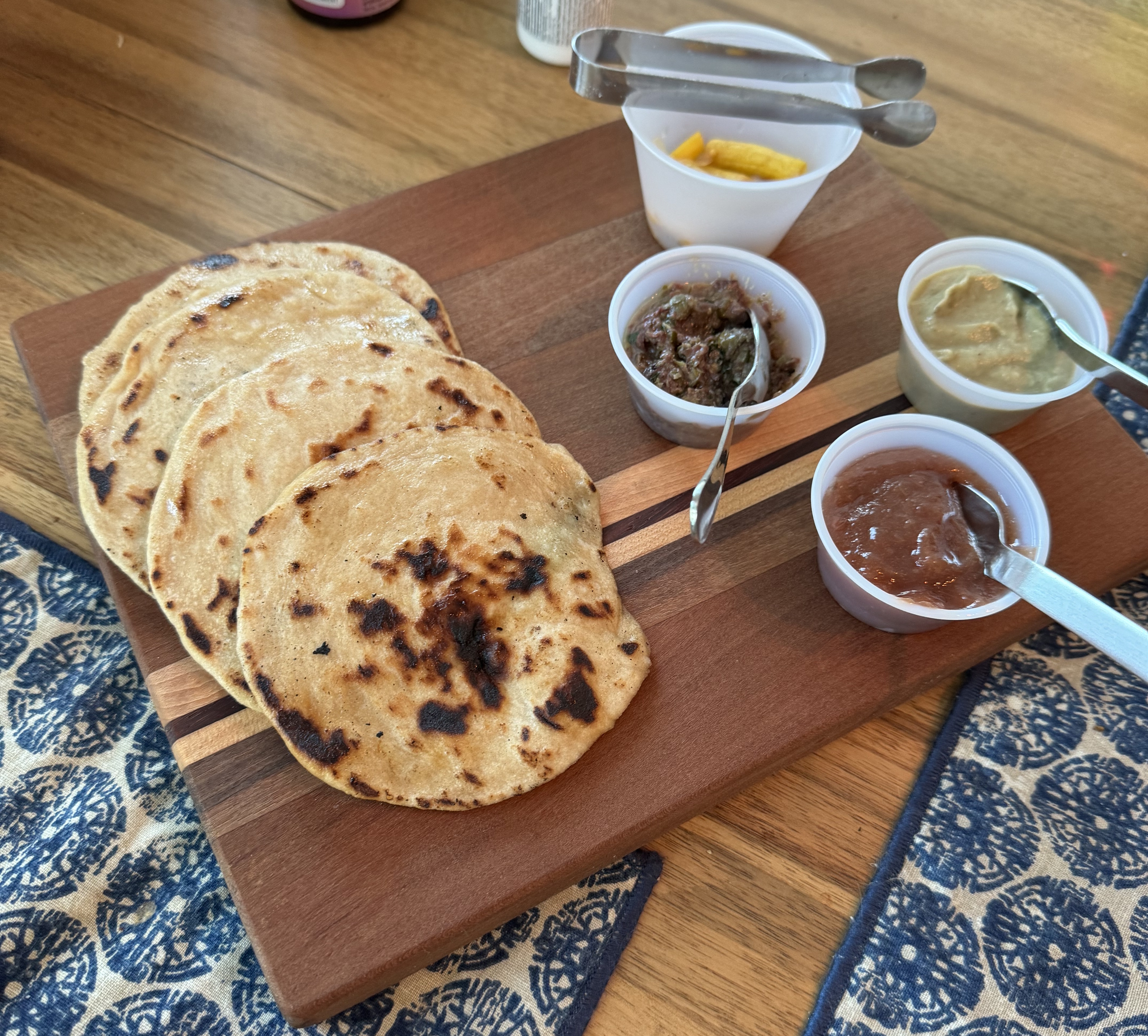 Stack of charred pita bread next to several small cups with dips and accoutrements. A tall cup has small yellow squash sliced lengthwise. One cup has a dark brown chunky dip. Another cup has a super-smooth, light brown dip. The last cup has a brick-red paste.