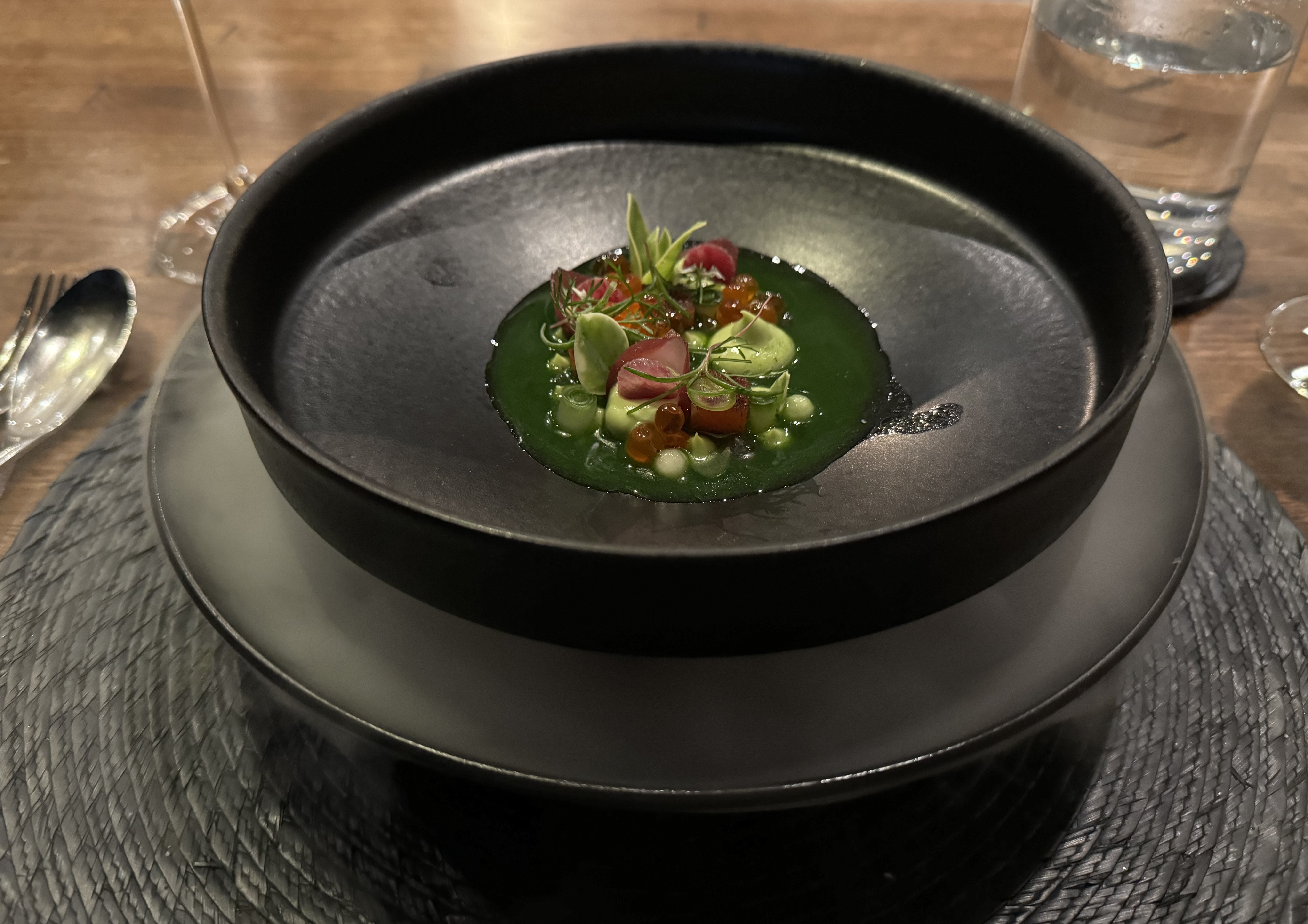 Two black bowls stacked up. The bottom bowl has generous amounts of fog pouring out of it. The top plate has a careful arrangement of salmon cubes, dollops of green sauce, and microgreen garnishes. The food is sitting in a pool of vibrant, dark green liquid