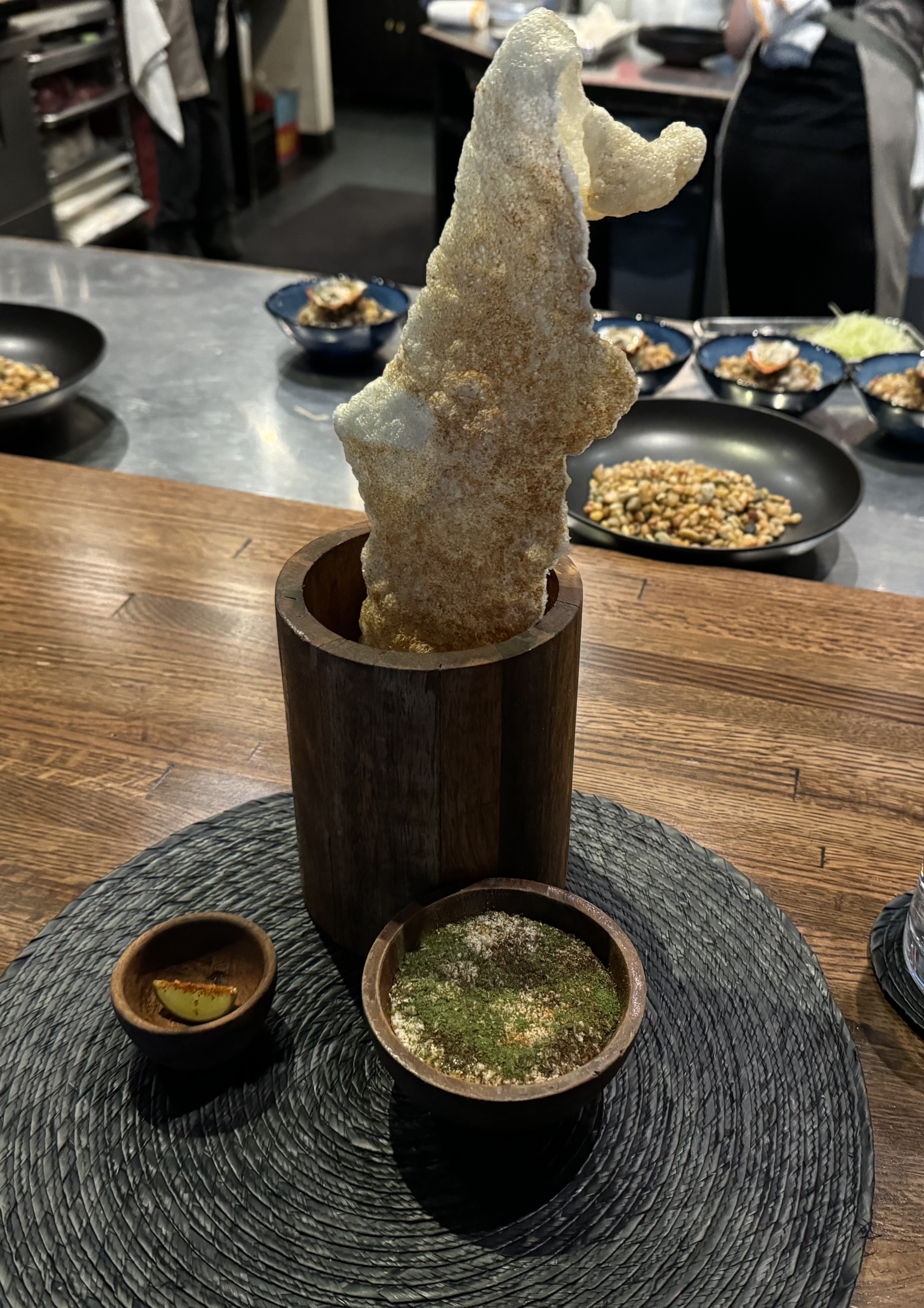 A tall wooden vase holding a massive piece of puffed skin, dusted with something red. Next to it is a small wooden bowl with a sauce dusted over with green powder, and a tiny wooden bowl with a wedge of lime. The lime wedge has red powder along its point. The vase and bowls all match.