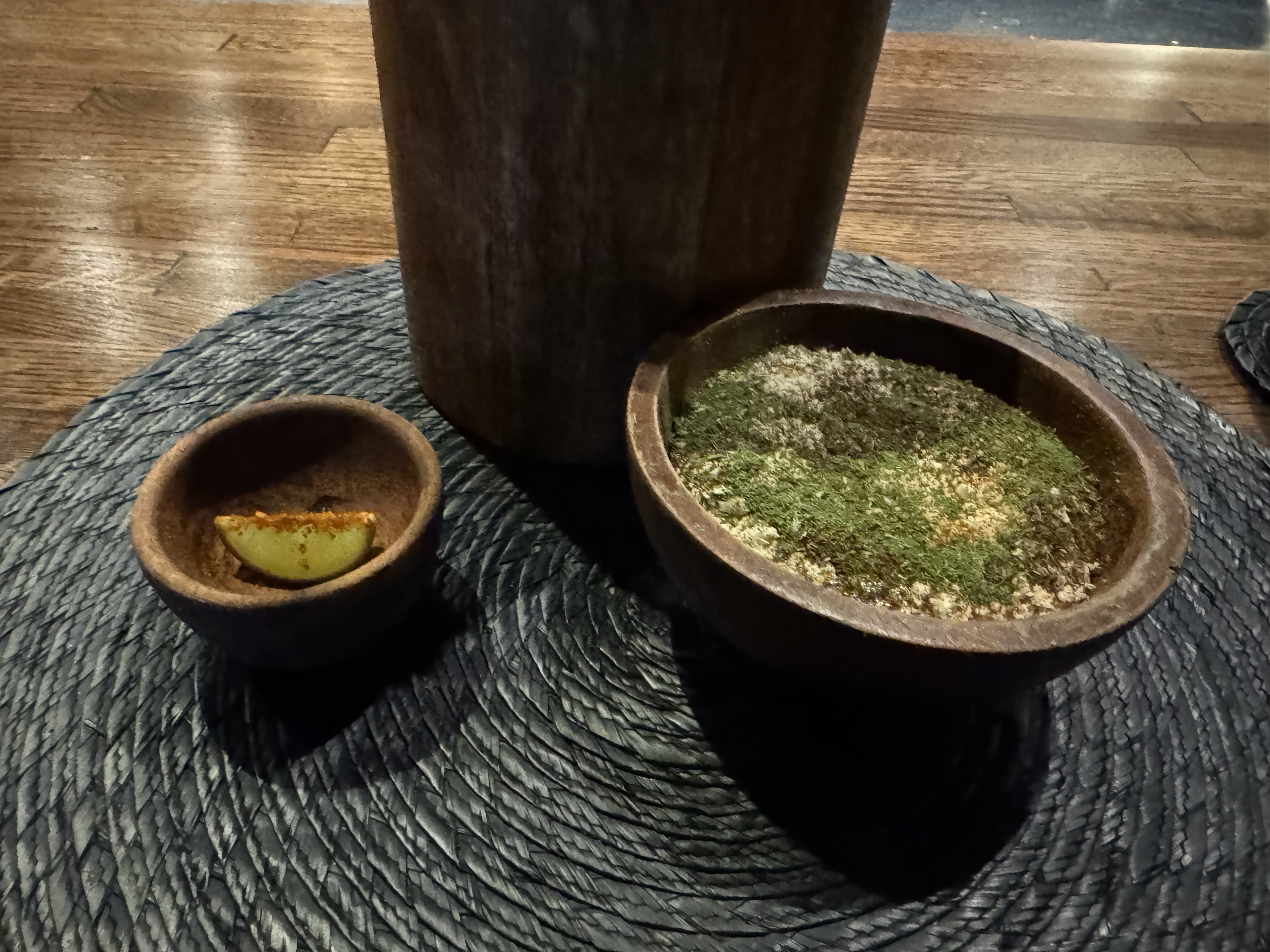 Close-up of the smaller wooden bowls next to the chicharrón. There is an off-white paste in one, covered in green dust. The smaller bowl has a lime wedge with a spicy powder on it.