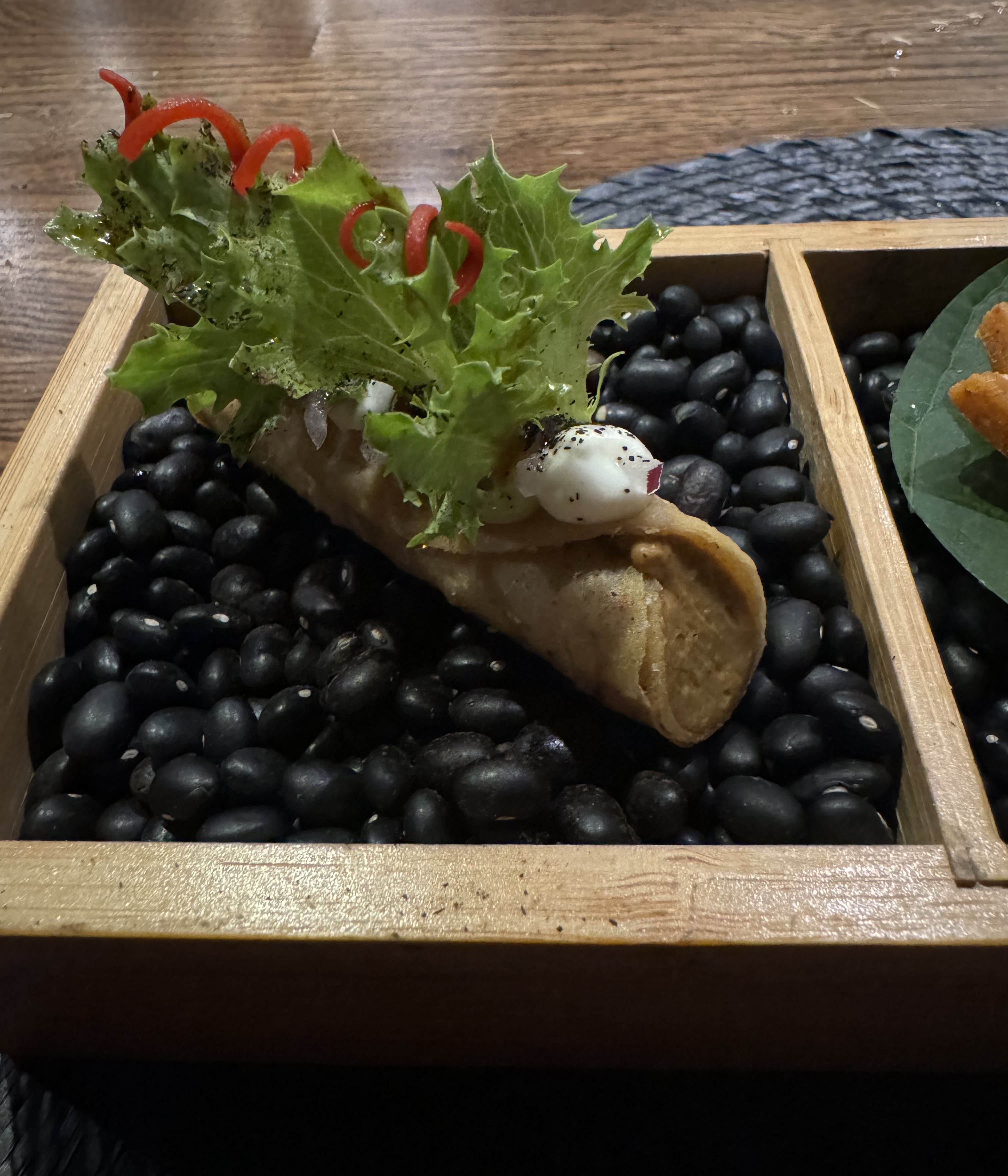 Close-up of one of the three small bites. This is a round shell filled with a pale brown mousse, much like a cannoli shell is filled with cheese. The are dollops of sour cream on top supporting some tiny lettuce leaves and curls of red pepper.