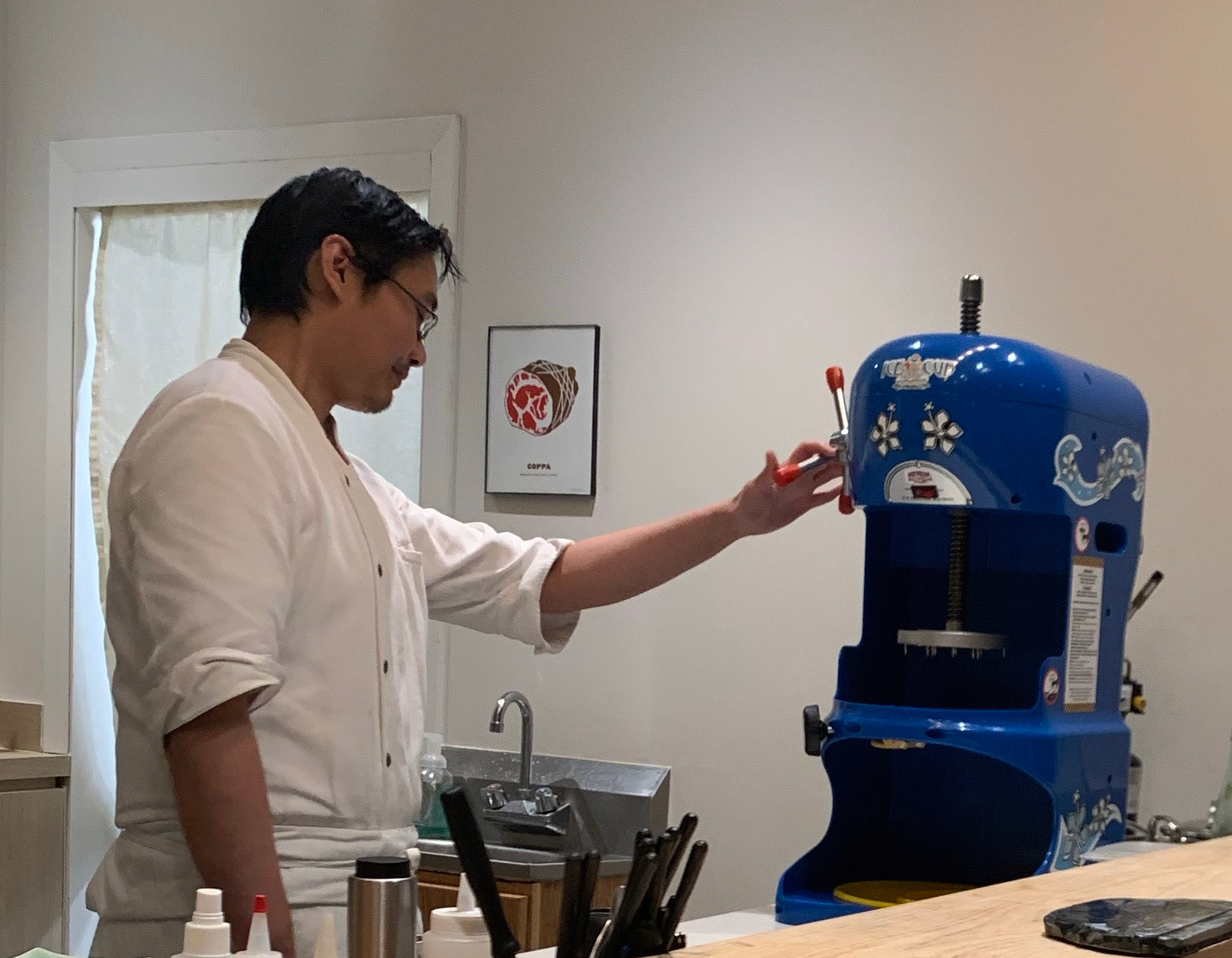 Man wearing glasses and chef attire standing next to a large blue machine. The machine has levers that control a disc. There is nothing inside the machine to be shaved by the disc yet.
