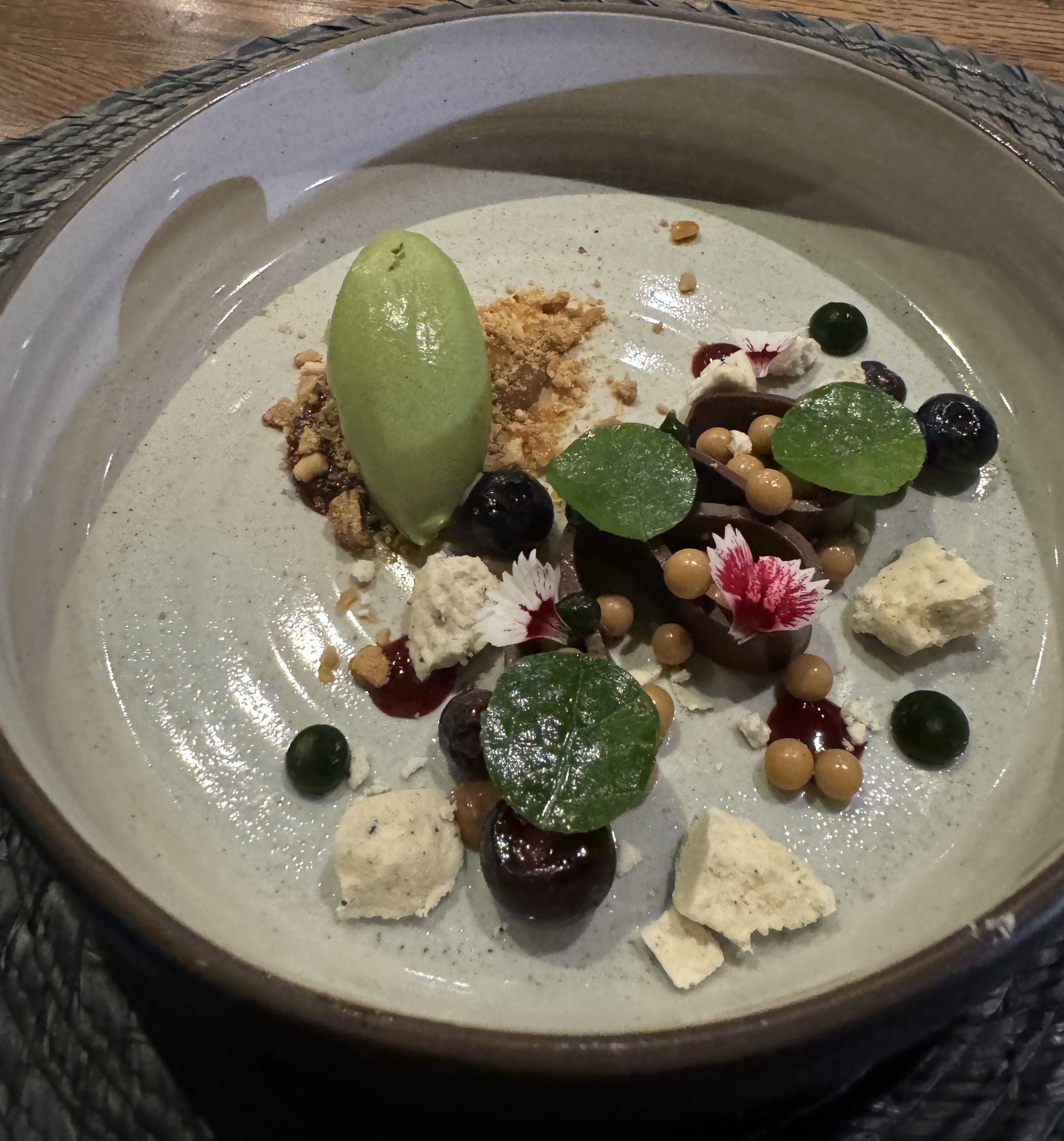 Shallow bowl with lots of ingredients carefully placed to look haphazardly scattered. There is a quenelle of green ice cream atop a crumble, bits of white meringue cookie, beads of light-brown, dots of black sauce, and shiny glazed green leaves. There is a ribbon of chocolate on its side, and many of the garnishes have nestled into its curves.