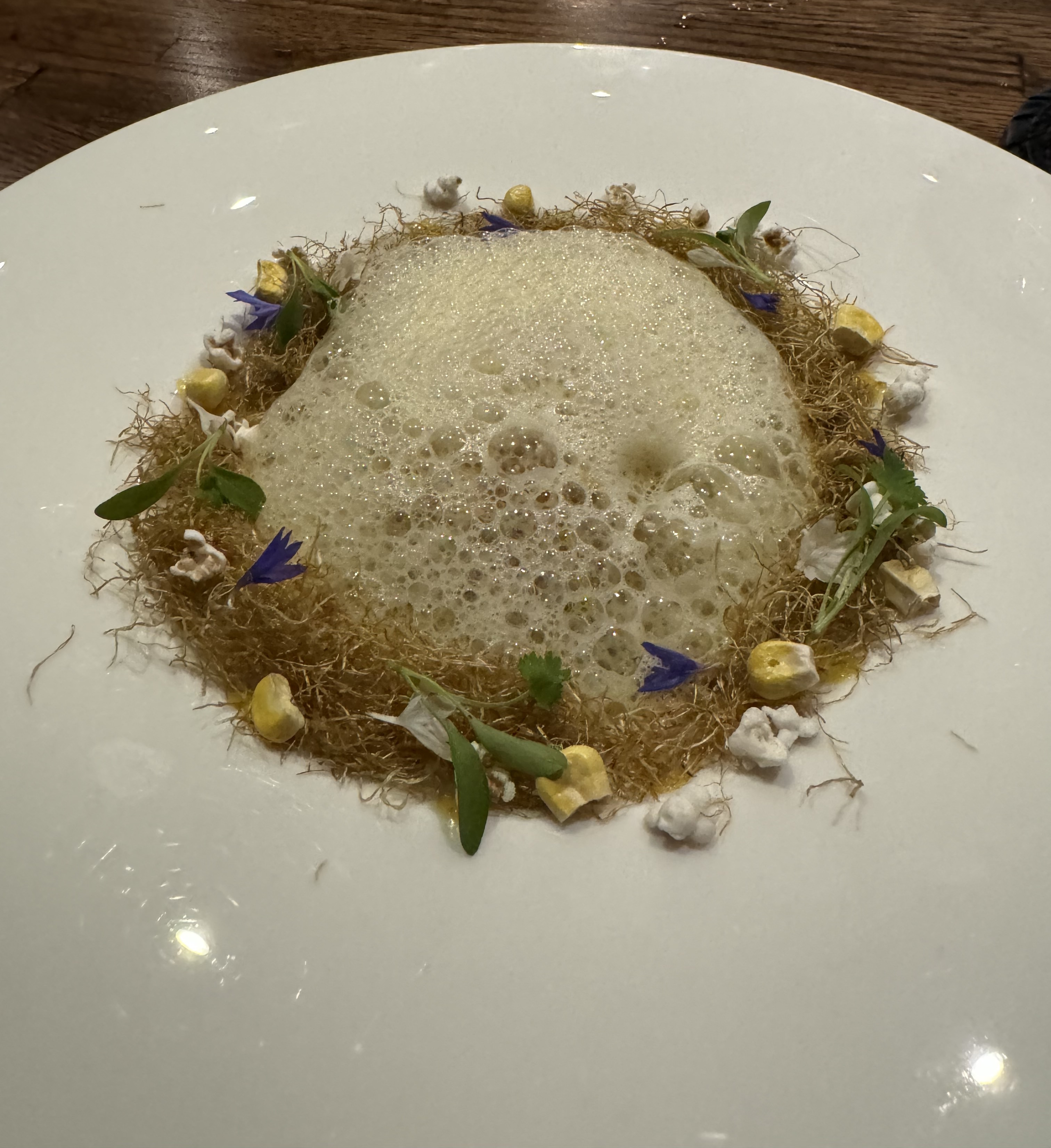 A very large round plate with a small dip in the center. It's filled with an off-white foam ringed by dried corn silk, dried corn kernels, bits of popcorn, and corn flowers.