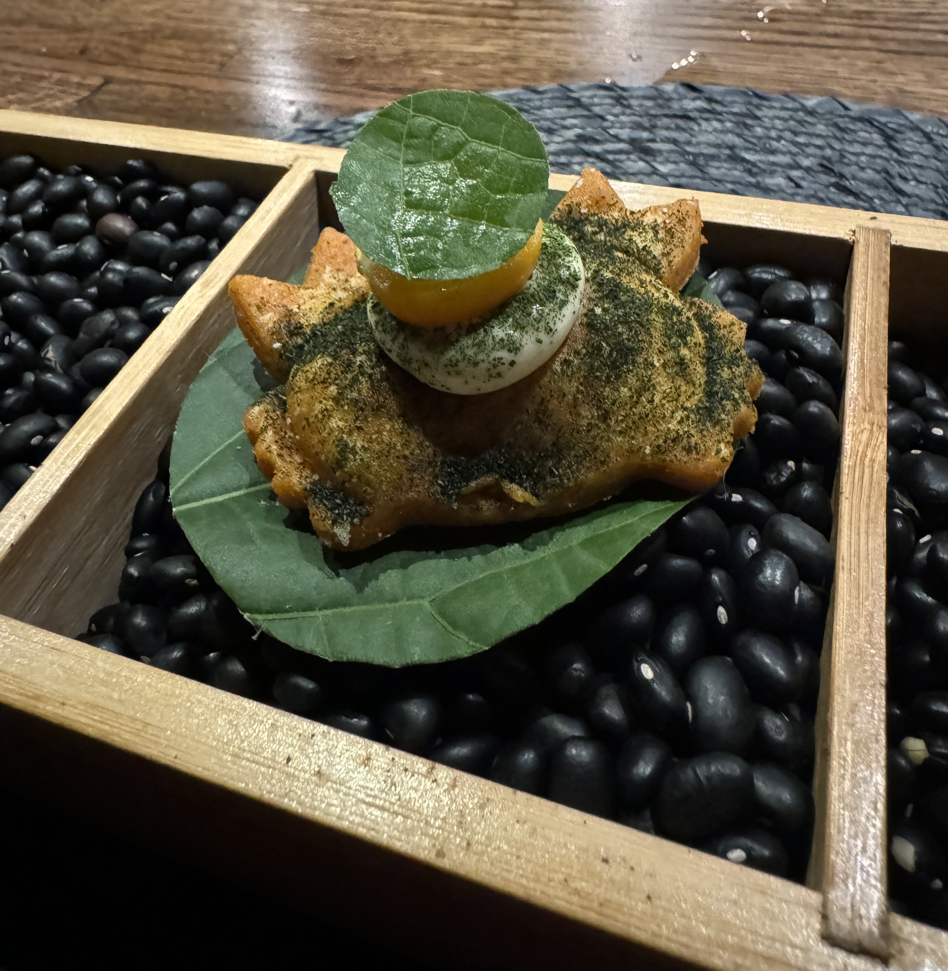 Crab-shaped empenada, complete with claws, sitting on top of a wide green leaf. The crab is dusted in green powder. It has a dollop of sour cream on top supporting a gooseberry and a smaller version of the green leaf it's sitting upon.