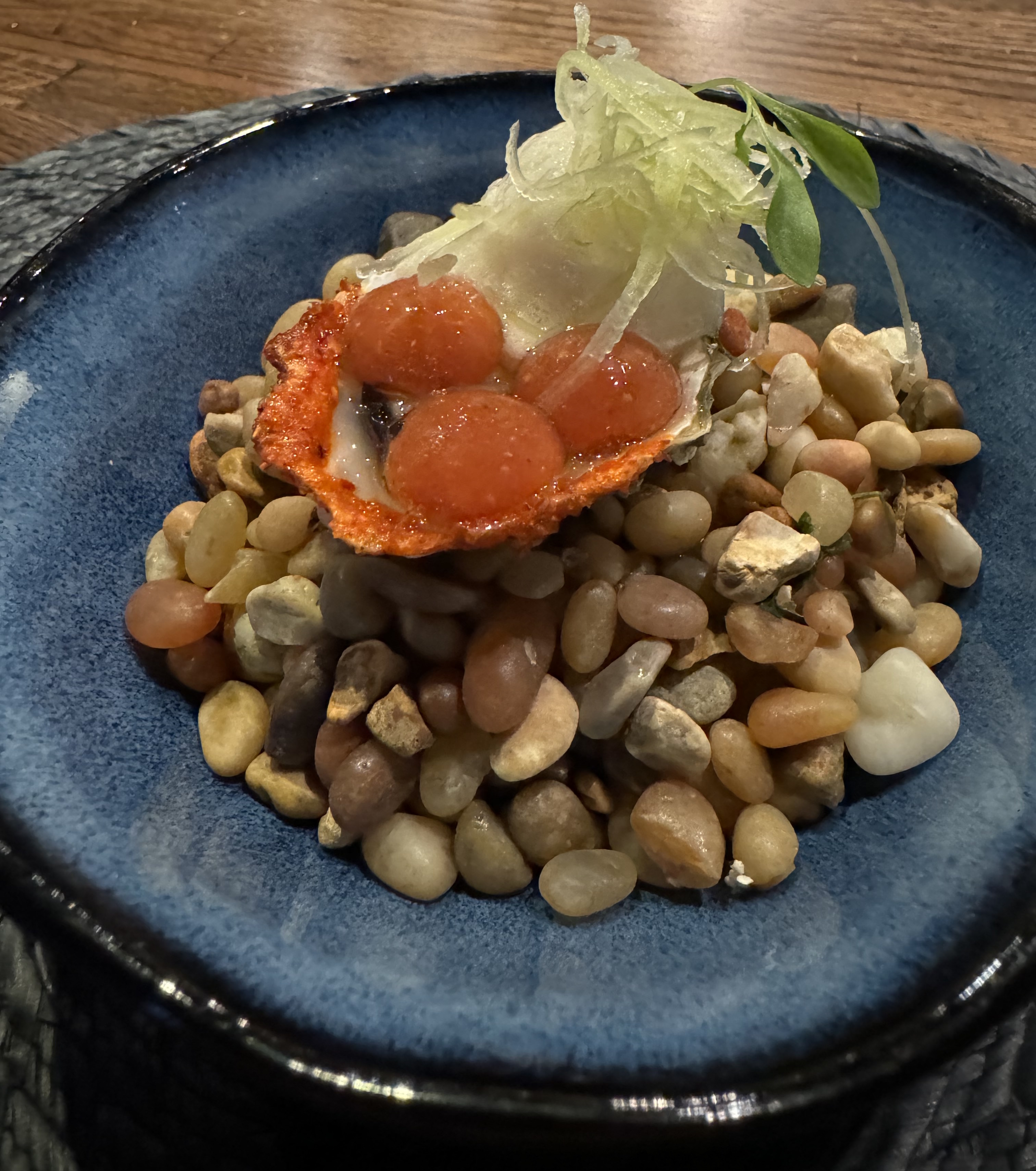 A fresh oyster still in its shell sitting atop a pile of multi-coloured rocks. The rocks are all rounded and smooth like driftwood. The oyster is rimmed with red spices. There are three bright-red orbs sitting atop the oyster meat, and some fluffy white foam behind them. The foam is topped with thin-shaved curls of a light green pepper.