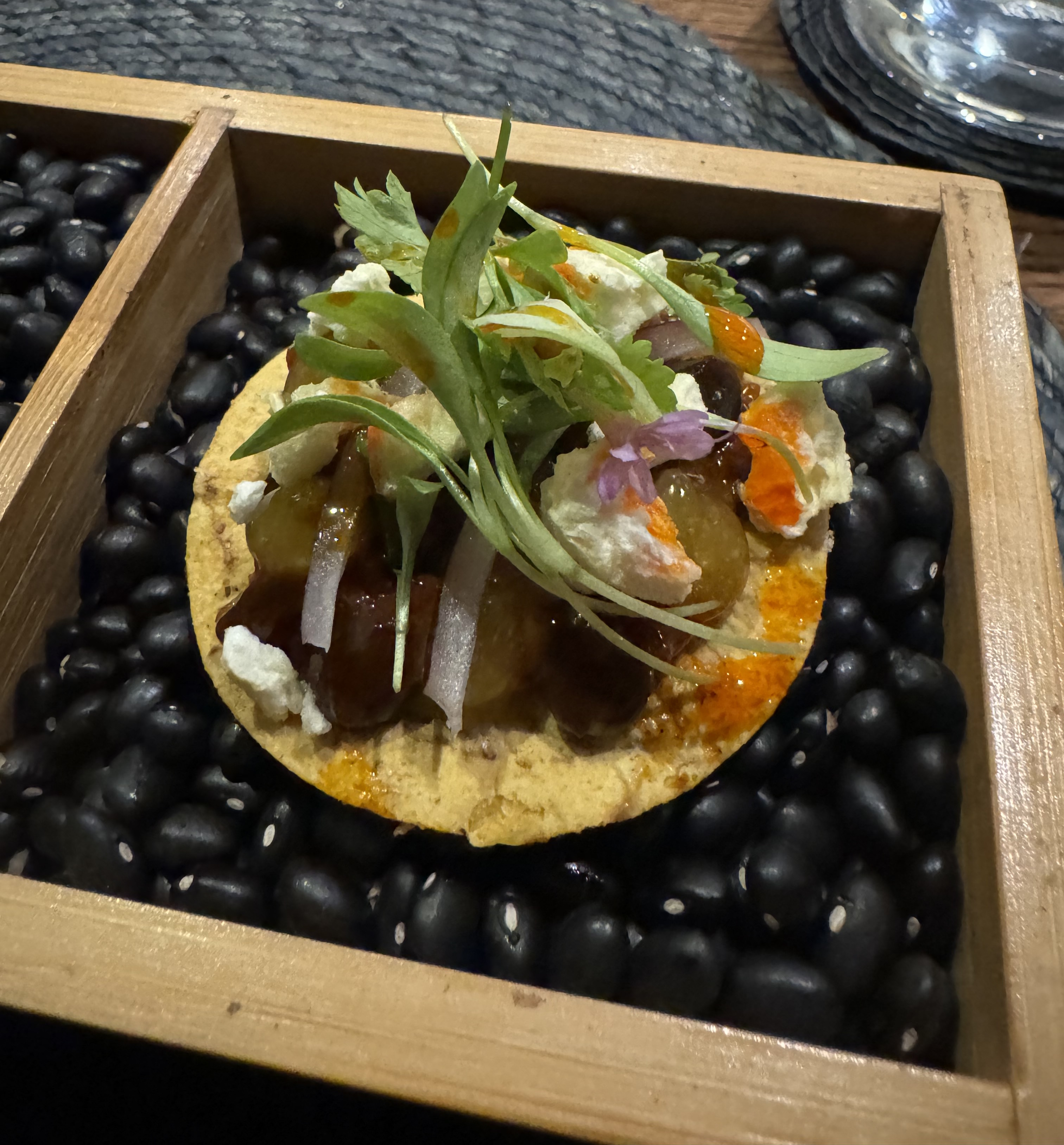 A round, yellow, crisp tortilla base supporting raw lamb cubes, a bright-red sauce, bits of cheese, pinapple, and some fresh greens for garnish