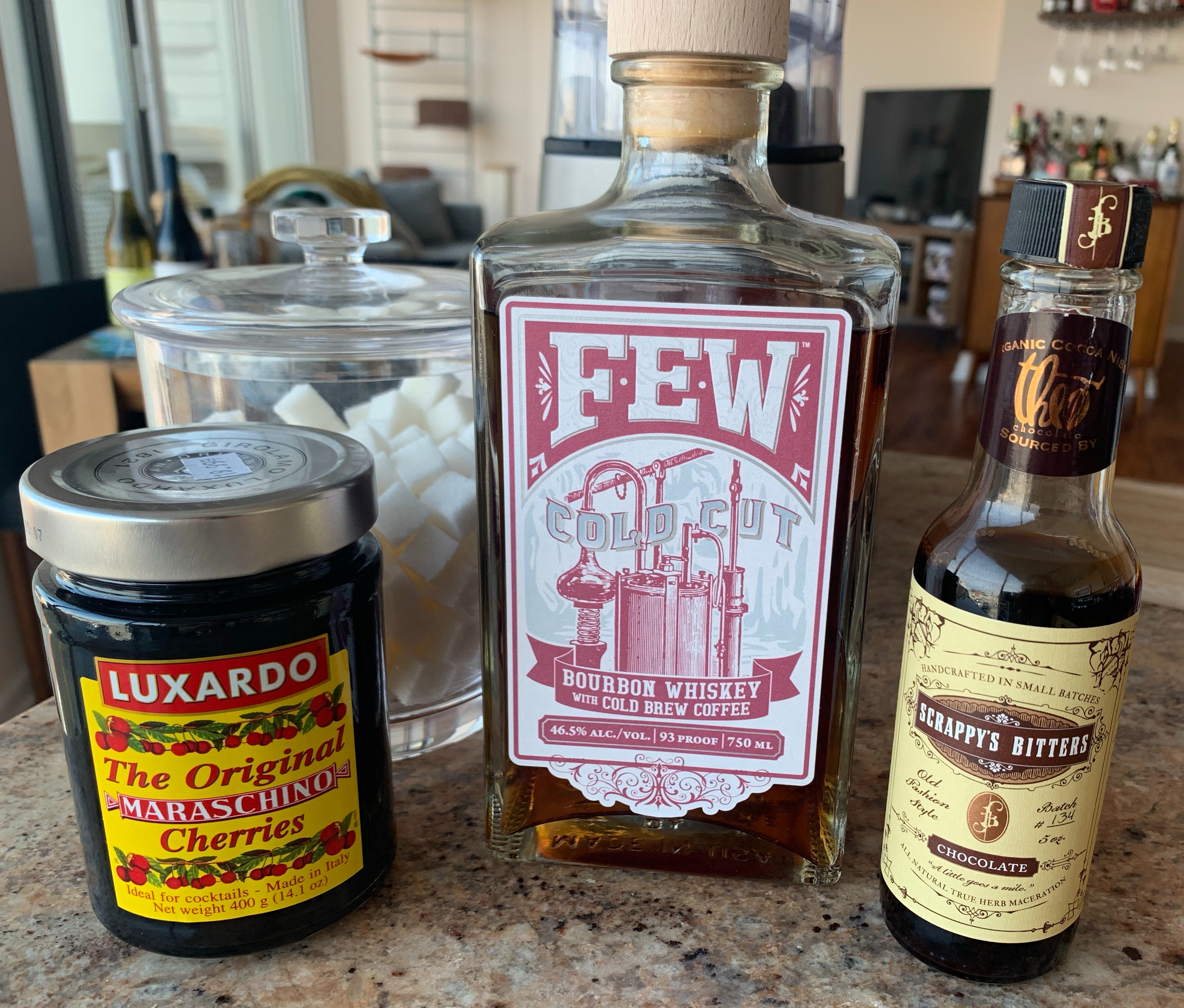 Bottle of the FEW Cold Cut Bourbon Whiskey with Cold Brew Coffee, flanked by chocolate bitters and a jar of maraschino cherries. A container of sugar cubes are in the background.