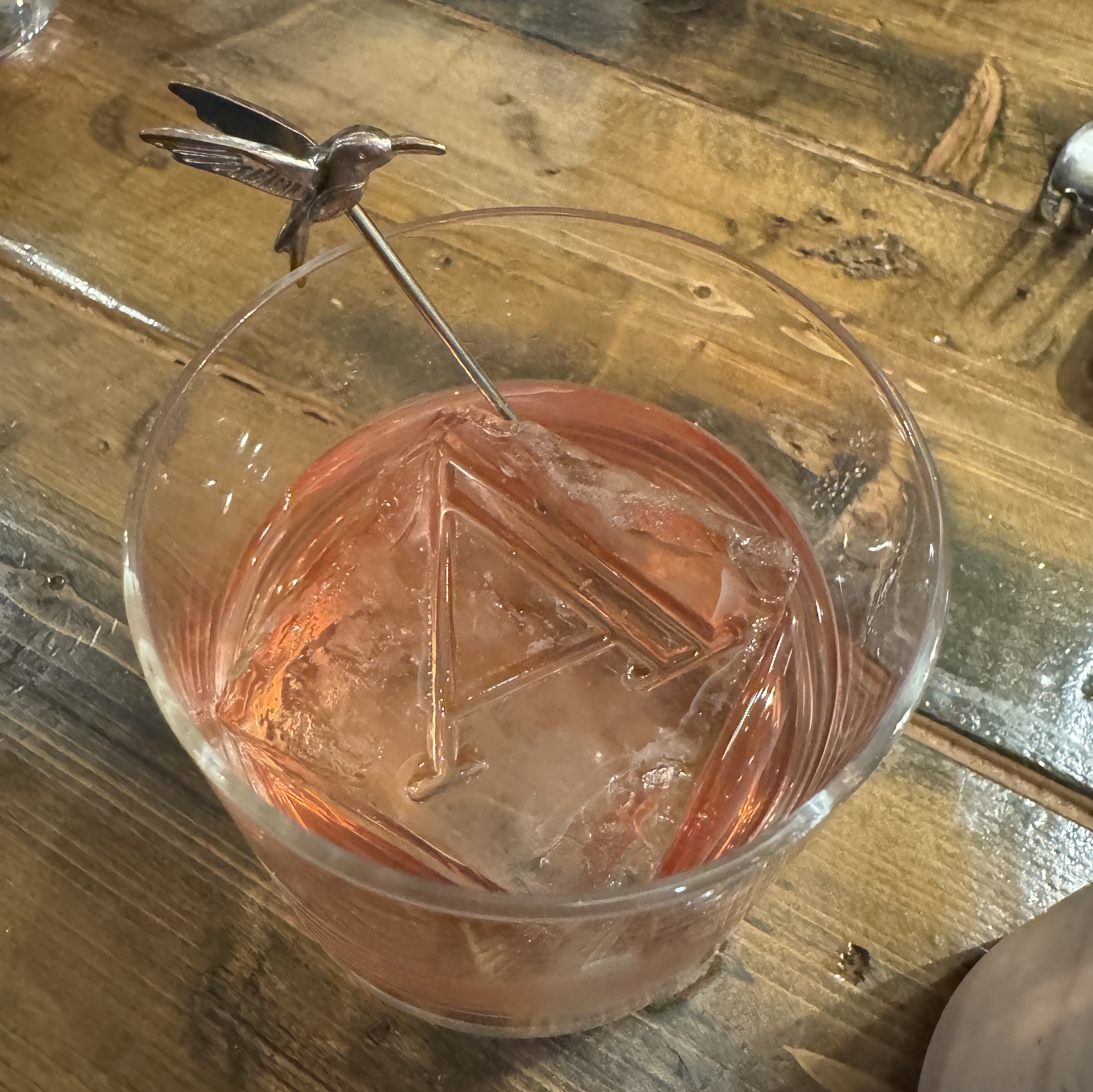 Pale pink fluid in a rocks glass. There's a huge cube of clear ice with the Atelier "A" logo melted into it, and a metal stirring rod topped by a hummingbird.