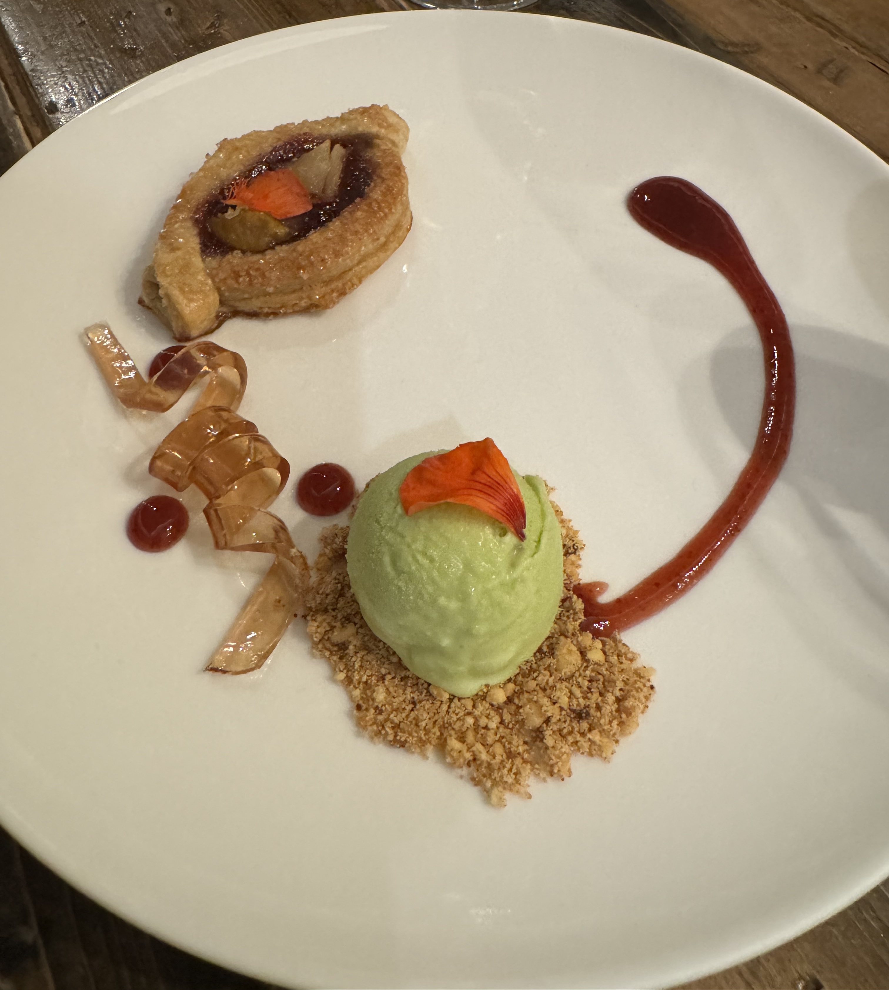 Plate with a golden-brown pastry, dusted in shimmering crystals of sugar, with some dark filling in the center. There are two shards of cooked pear emerging from the filling. Next to the pastry is a twist of jelly. Next to that is a dusting of brown-butter crumble and green ice cream. There's a thick red plum sauce around the edge of the plate.