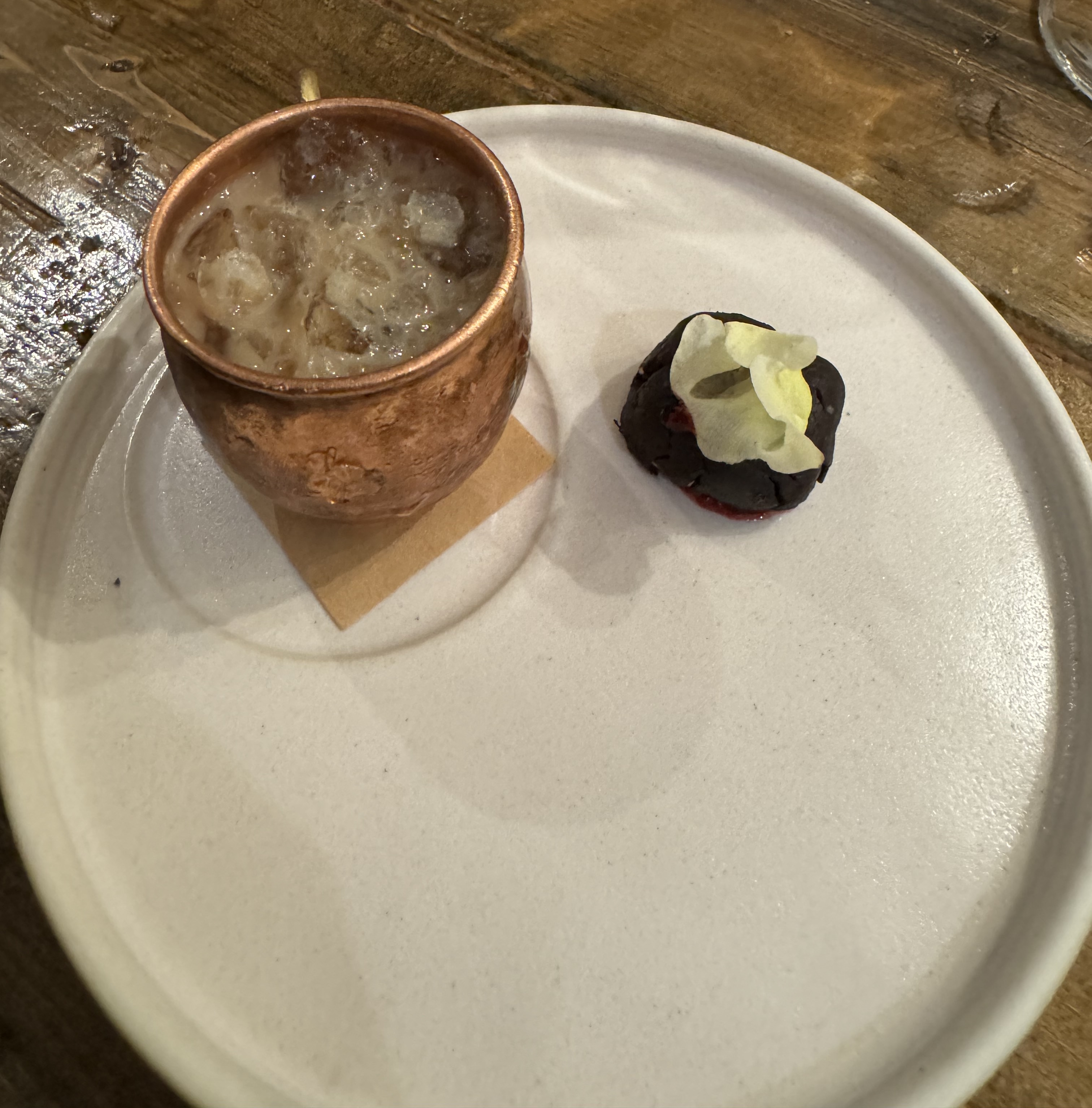 Large plate with two items upon it. One is a small jet-black cookie garnished with a flower. The other is a hammered copper mug with ice and milk.