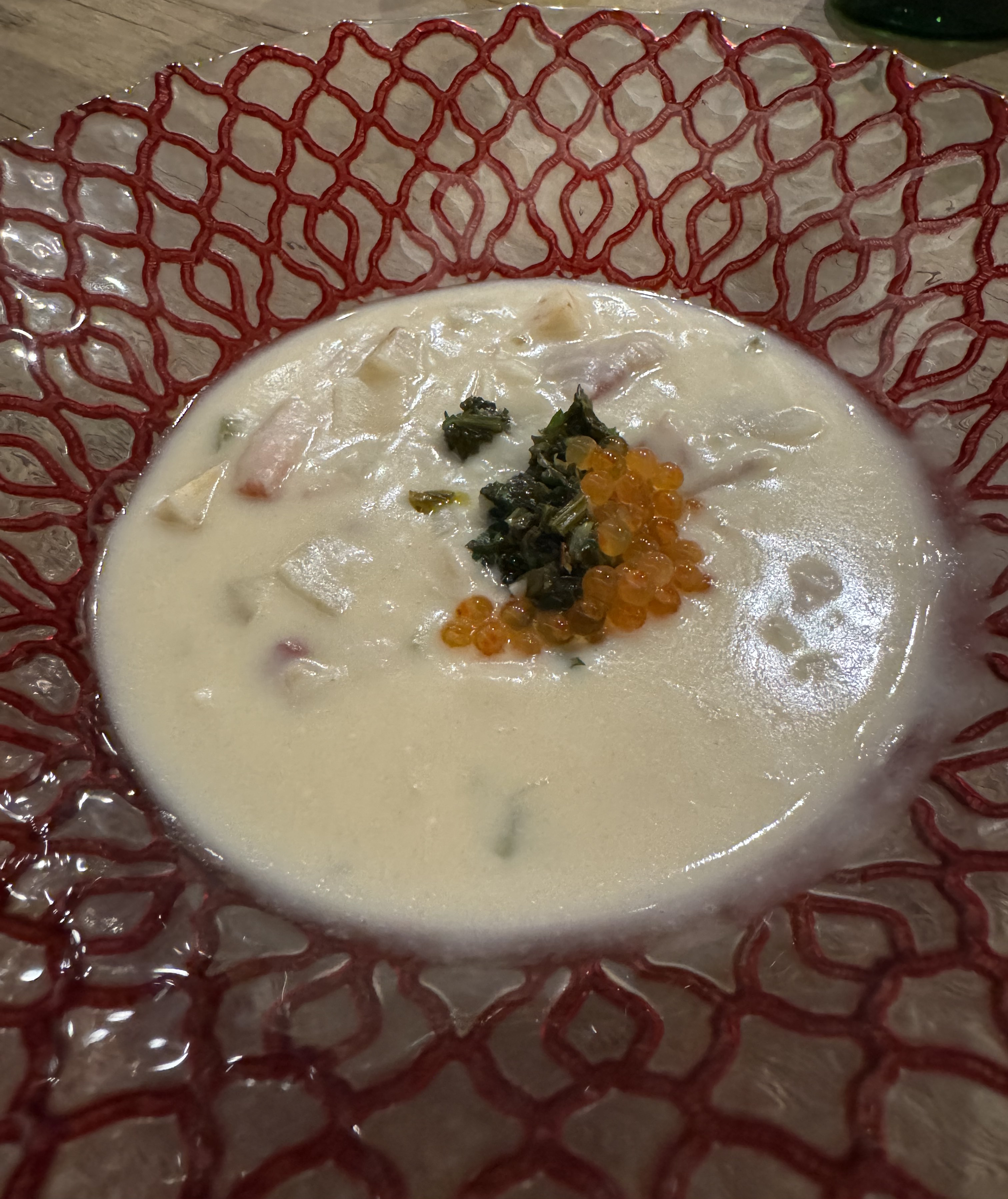 Bowl of white creamy soup. There are chunks barely visible just below the surface: cubes of pink and white. In the center, there's a small heap of a green sauce, and a bit of orange roe.